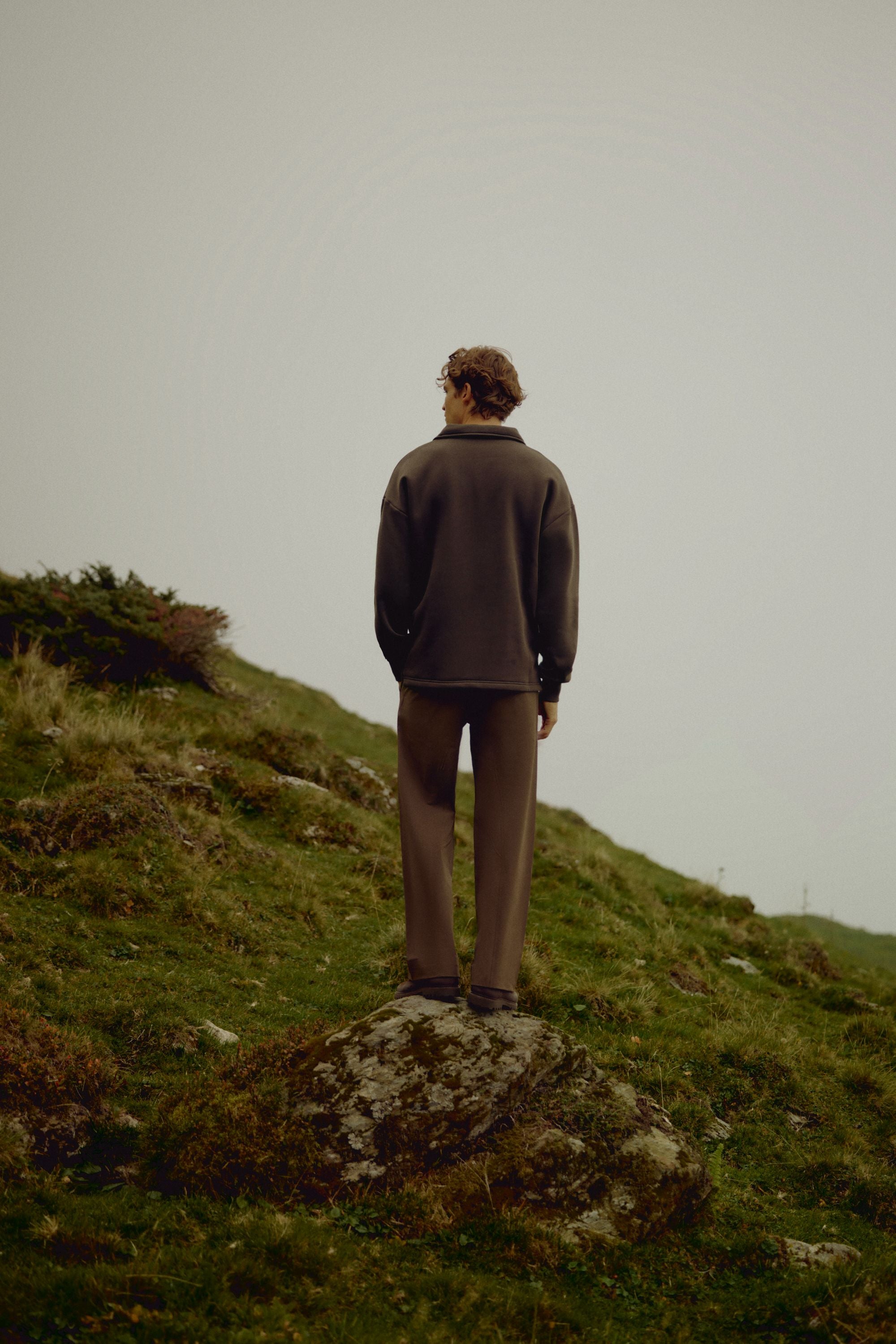 Man wearing an espresso CASPARA rugby shirt standing on a grassy hillside.