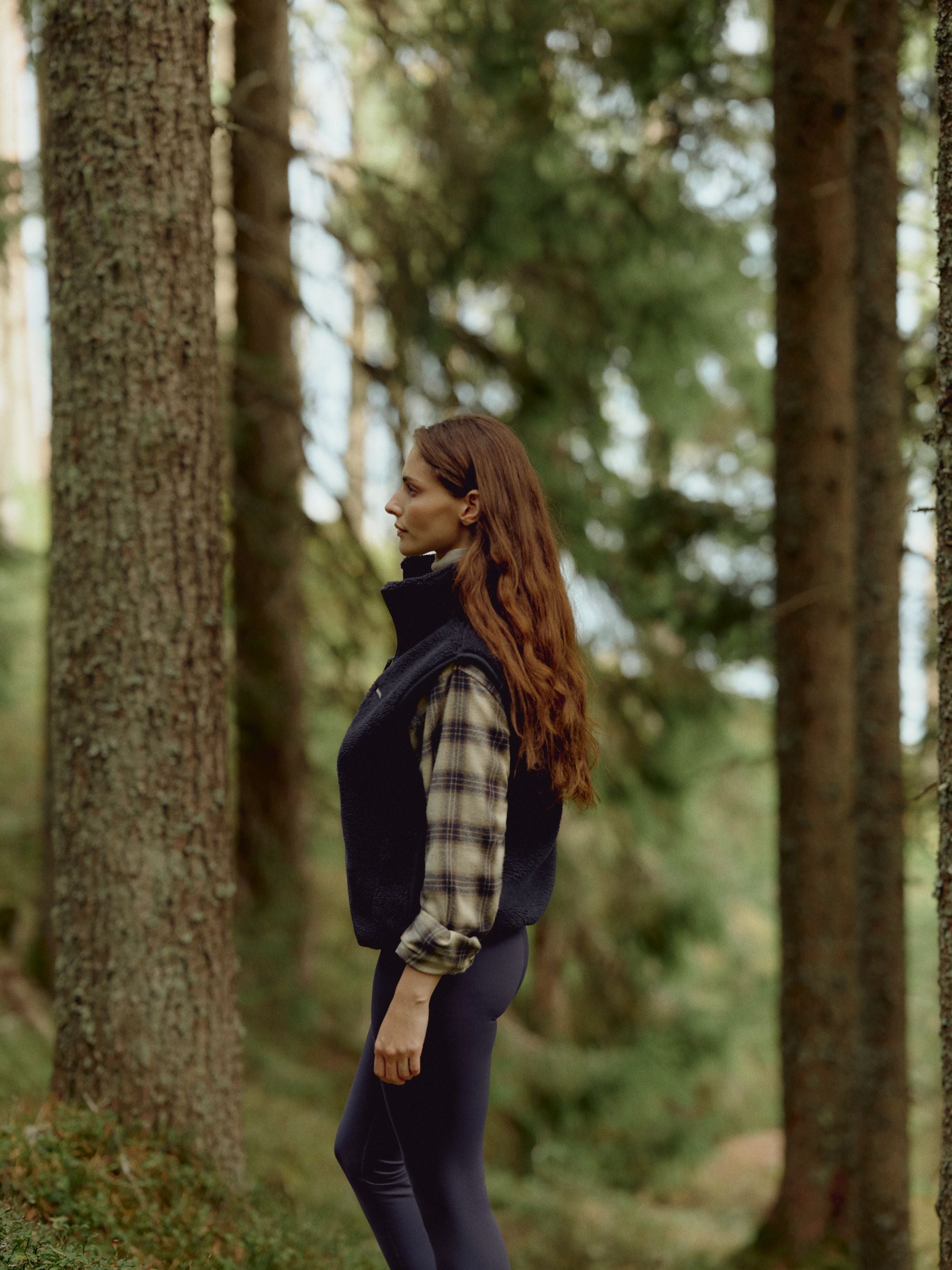 Woman wearing a navy CASPARA Cloudfleece vest standing in a pine forest.