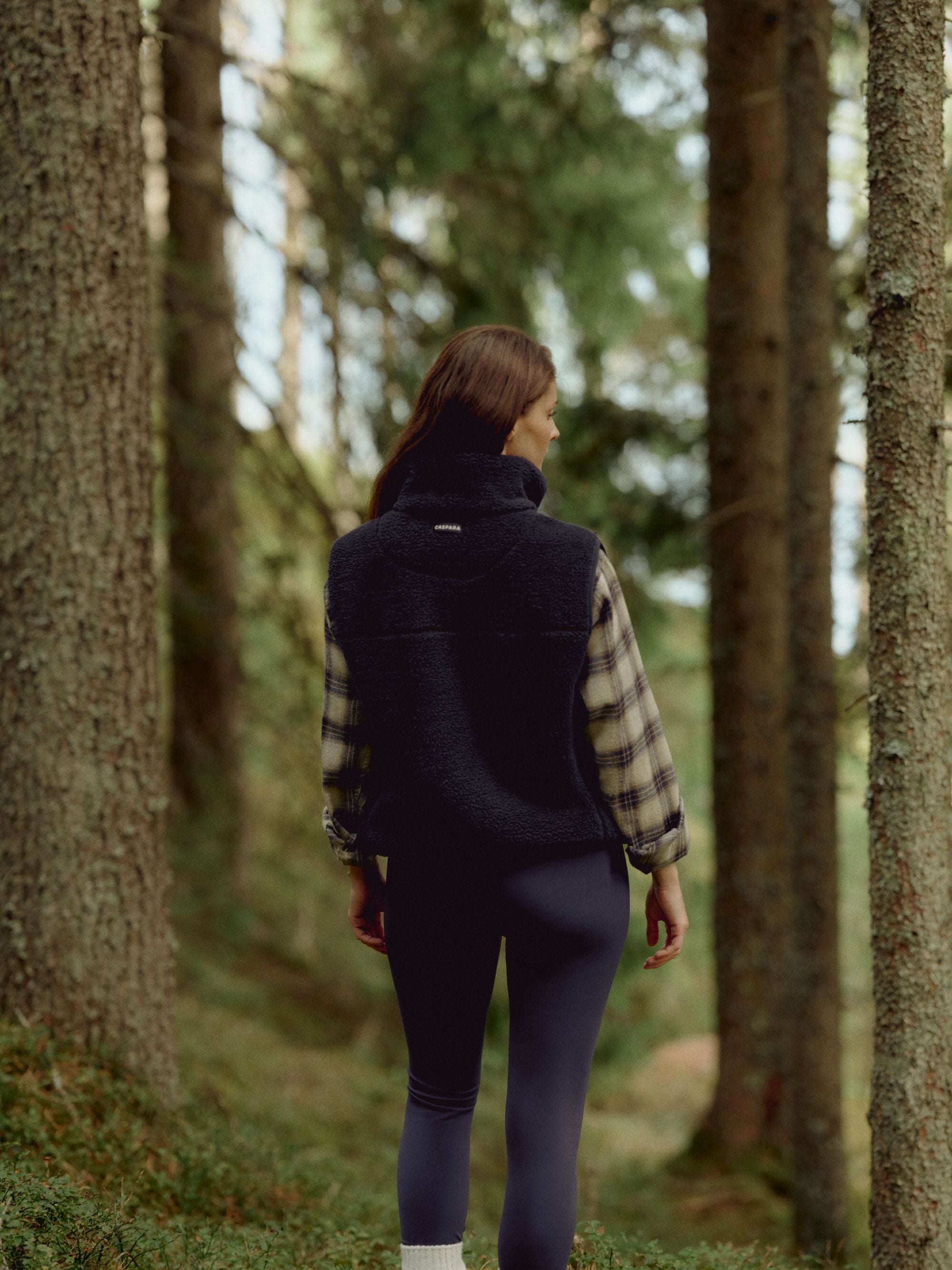 Woman wearing a navy CASPARA Cloudfleece vest in a pine forest, seen from behind.