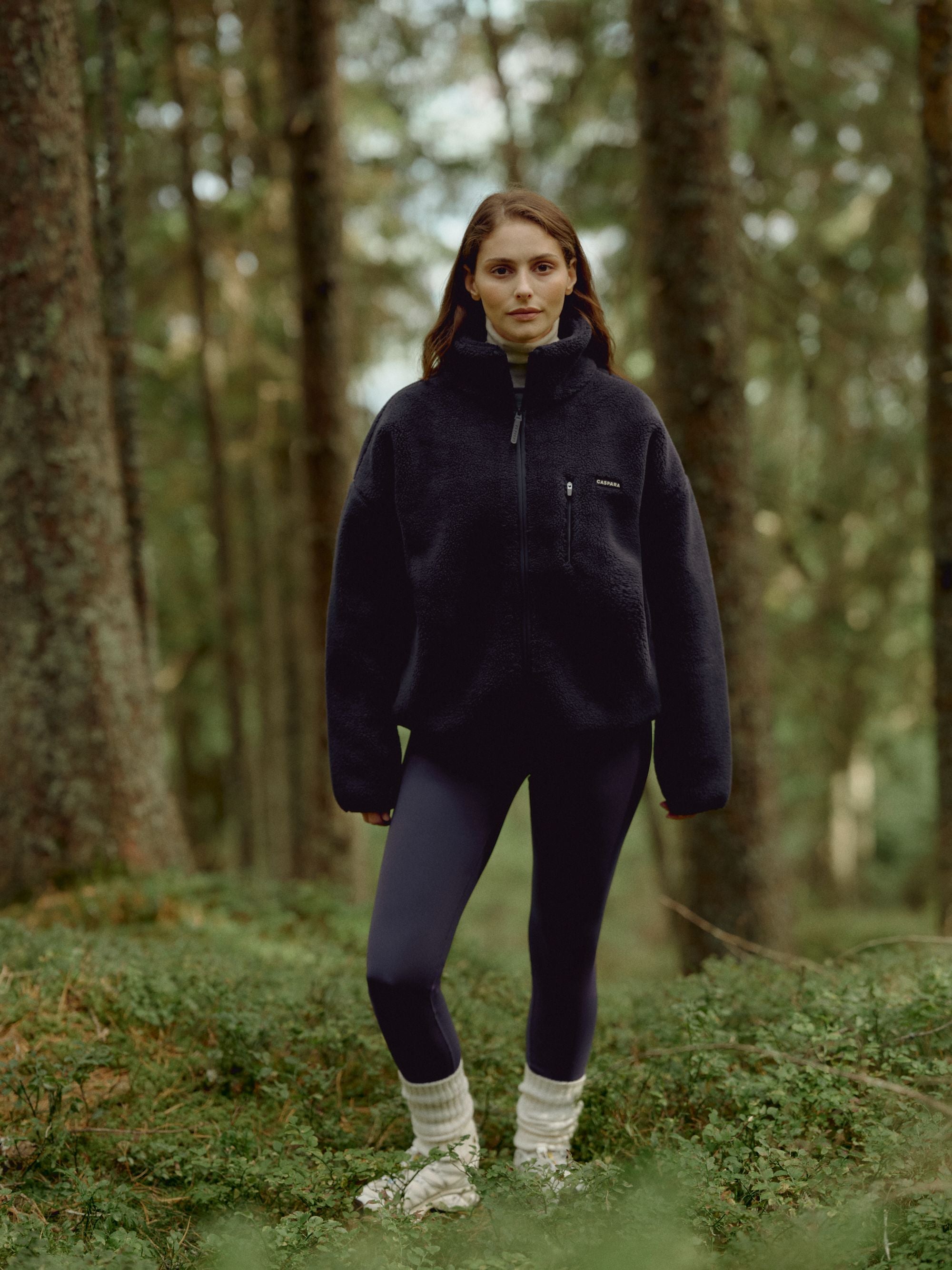 Woman wearing a navy CASPARA Cloudfleece jacket standing in a pine forest.