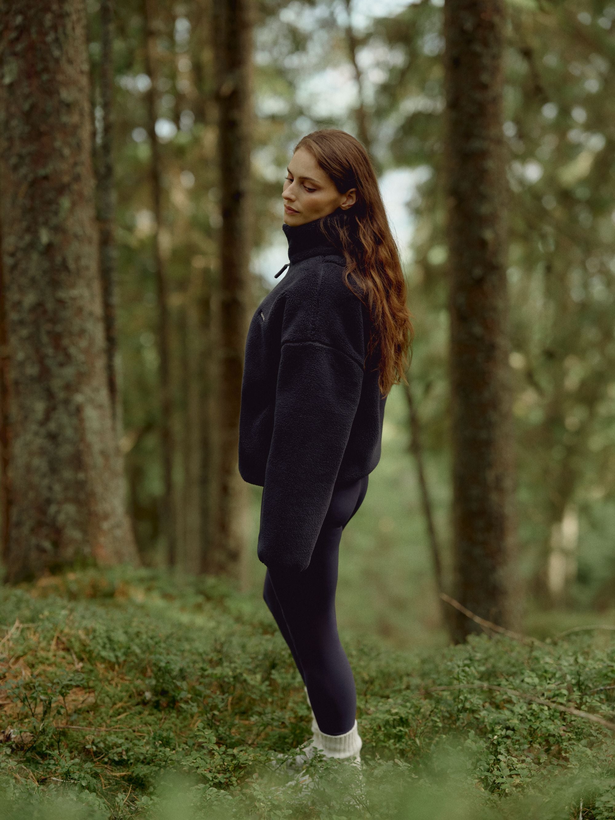 Woman wearing a navy CASPARA Cloudfleece jacket standing in a pine forest.