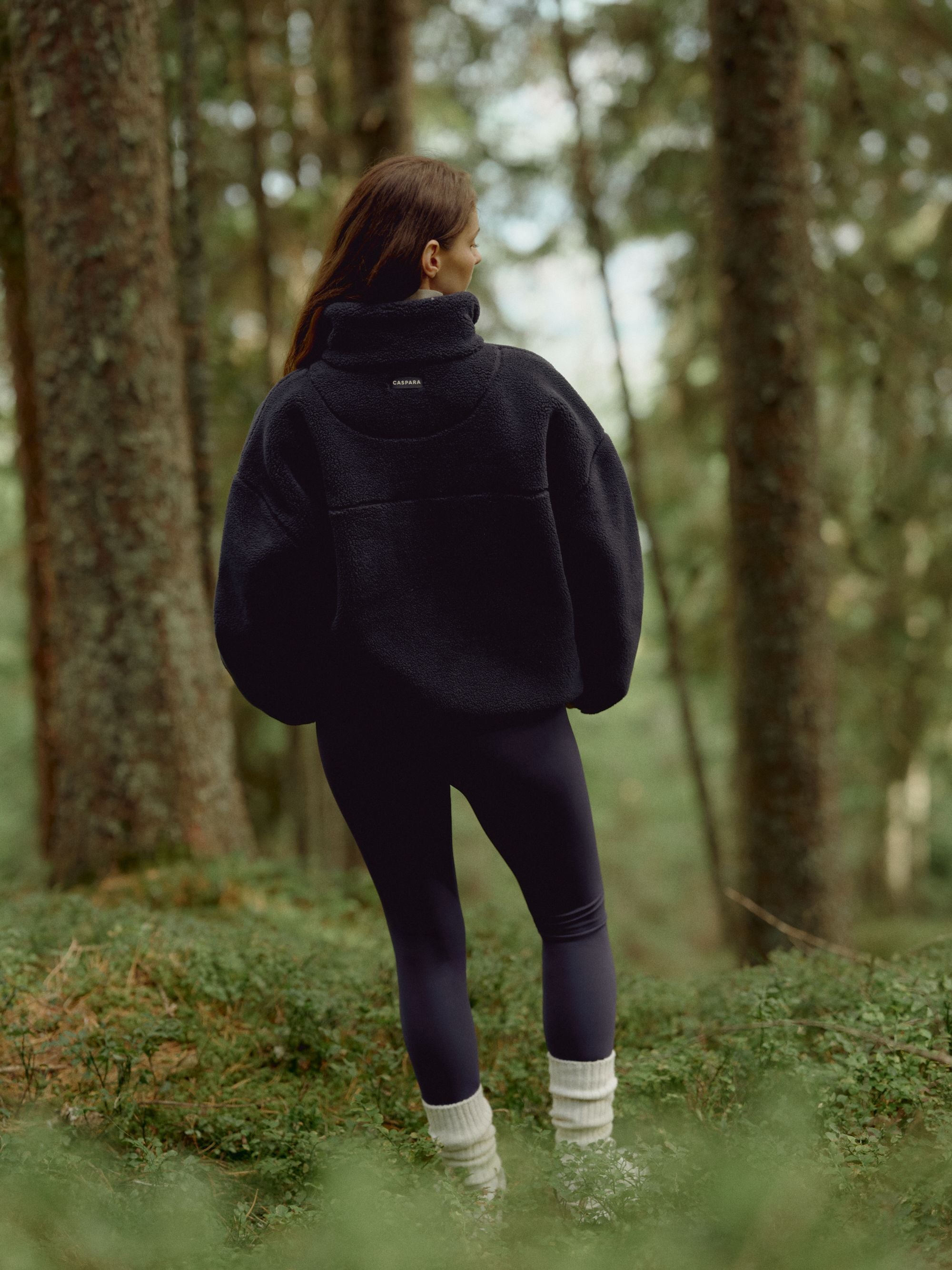 Woman wearing a navy CASPARA Cloudfleece jacket standing in a forest, seen from behind.