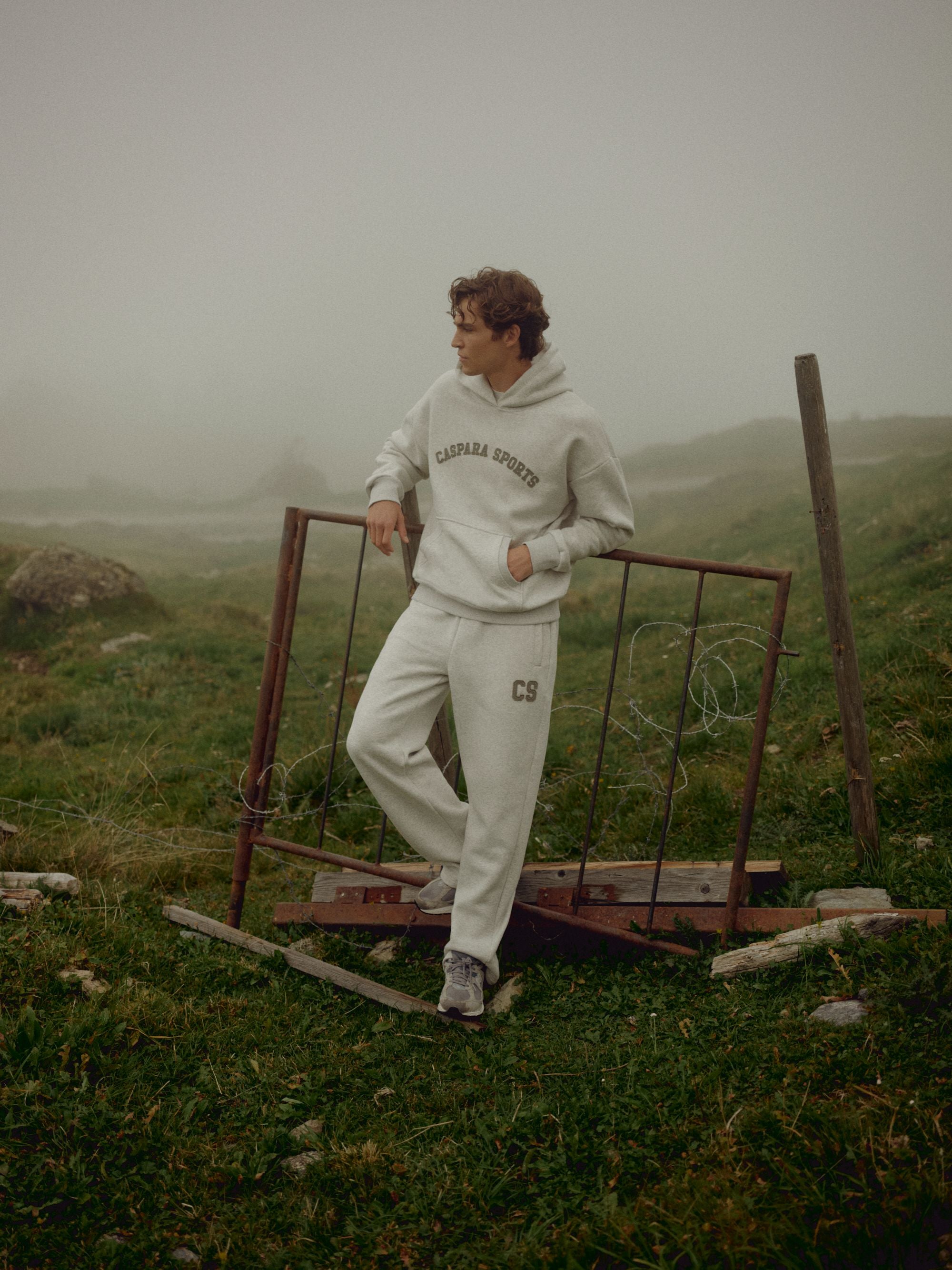 Man wearing a grey melange CASPARA CS hoodie leaning against a fence in a foggy landscape.