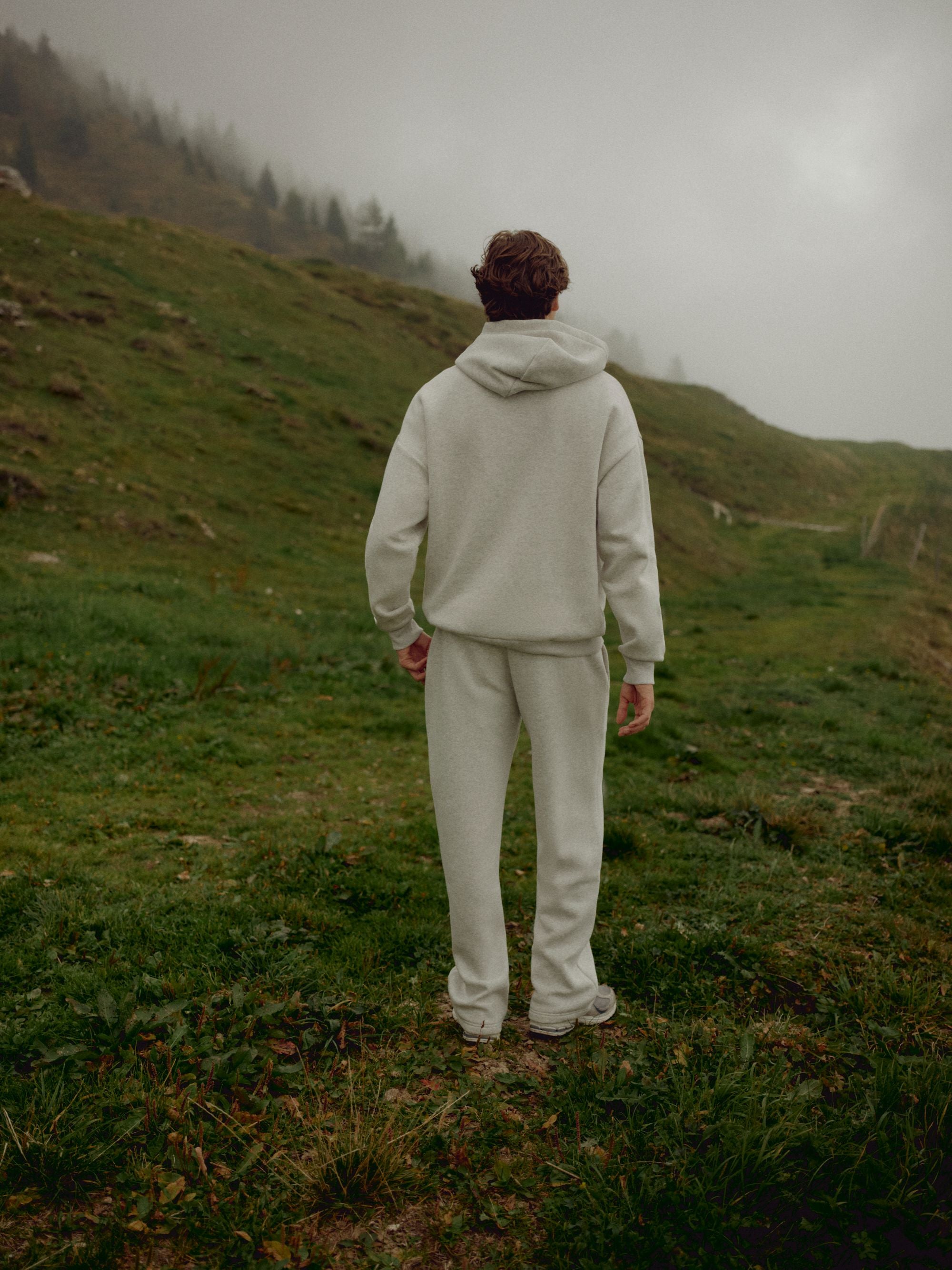 Man wearing a grey melange CASPARA CS hoodie standing on a foggy mountain hillside.