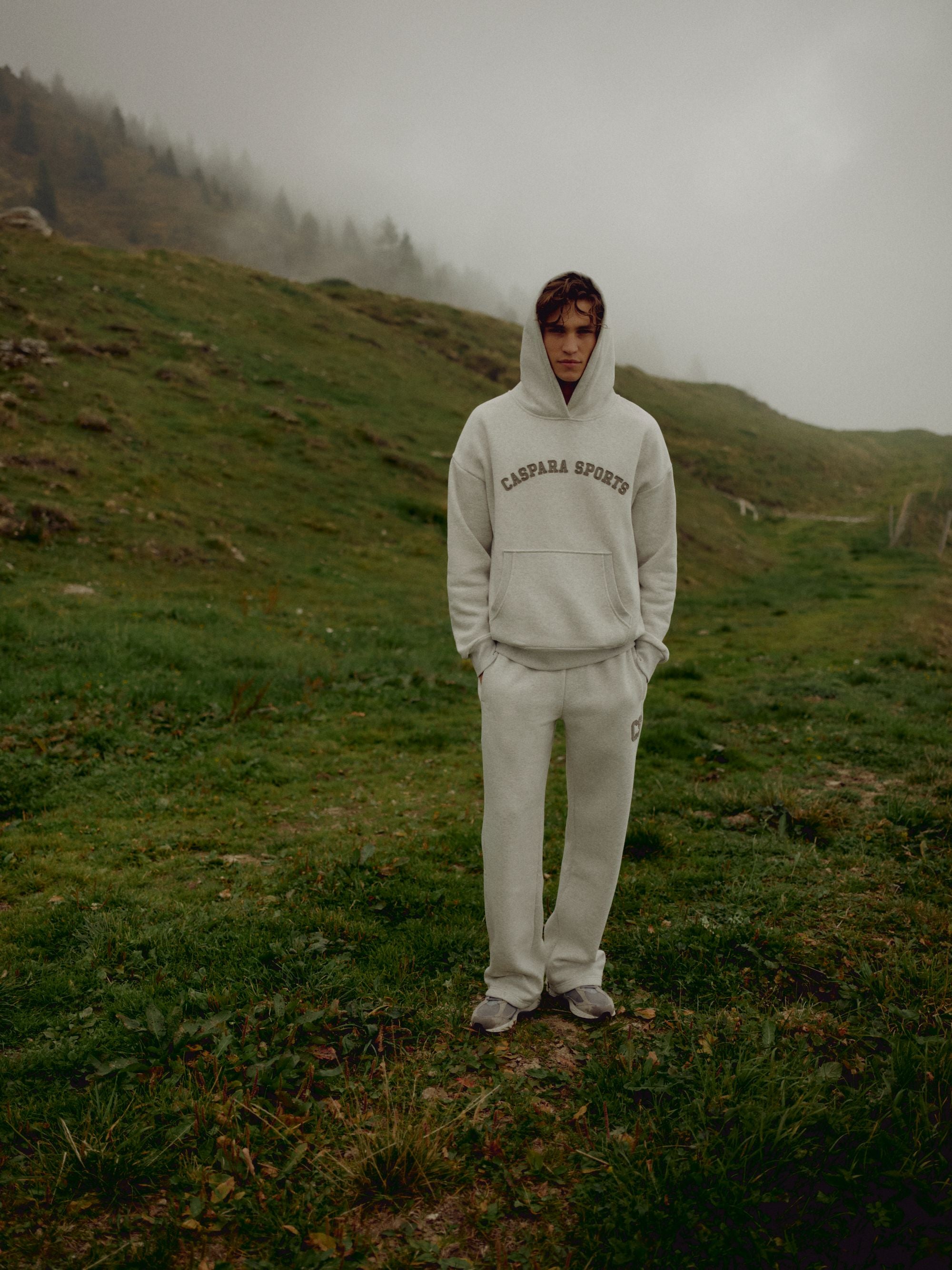 Man wearing a grey melange CASPARA CS hoodie standing on a misty hillside.