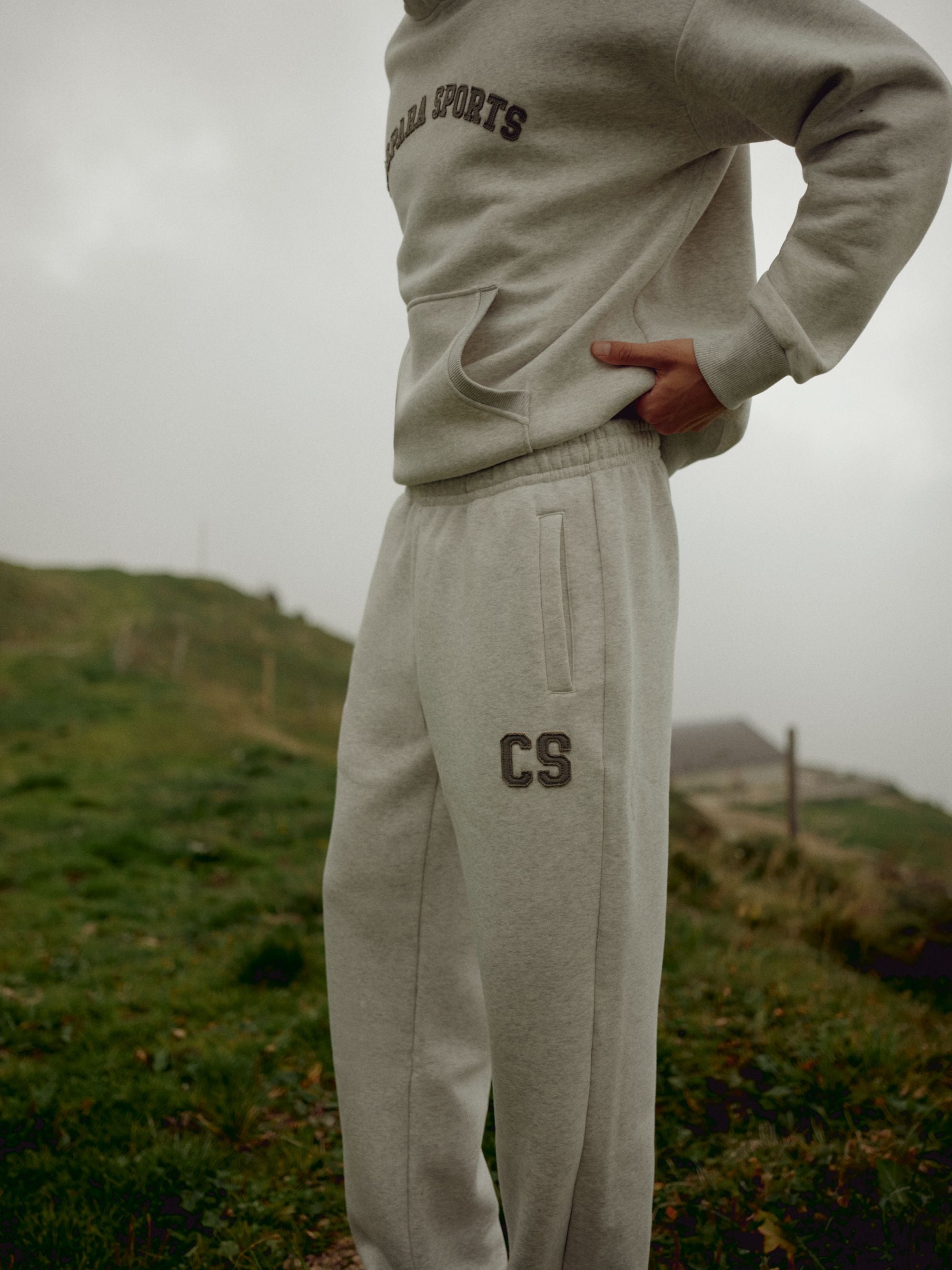 Man wearing a grey melange CASPARA CS hoodie and sweatpants on a grassy hillside.