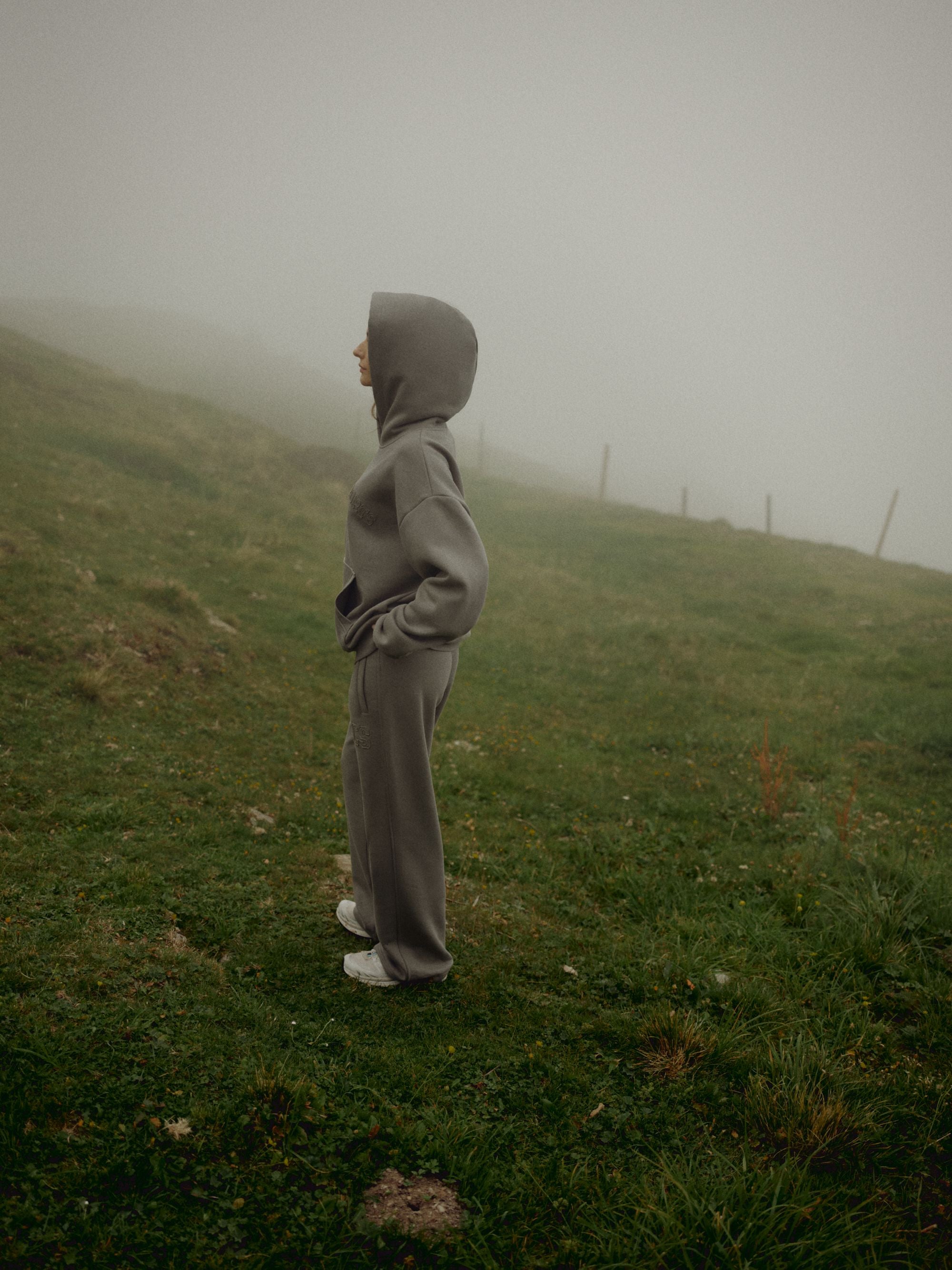 Woman wearing a khaki CASPARA CS hoodie standing on a foggy hillside.
