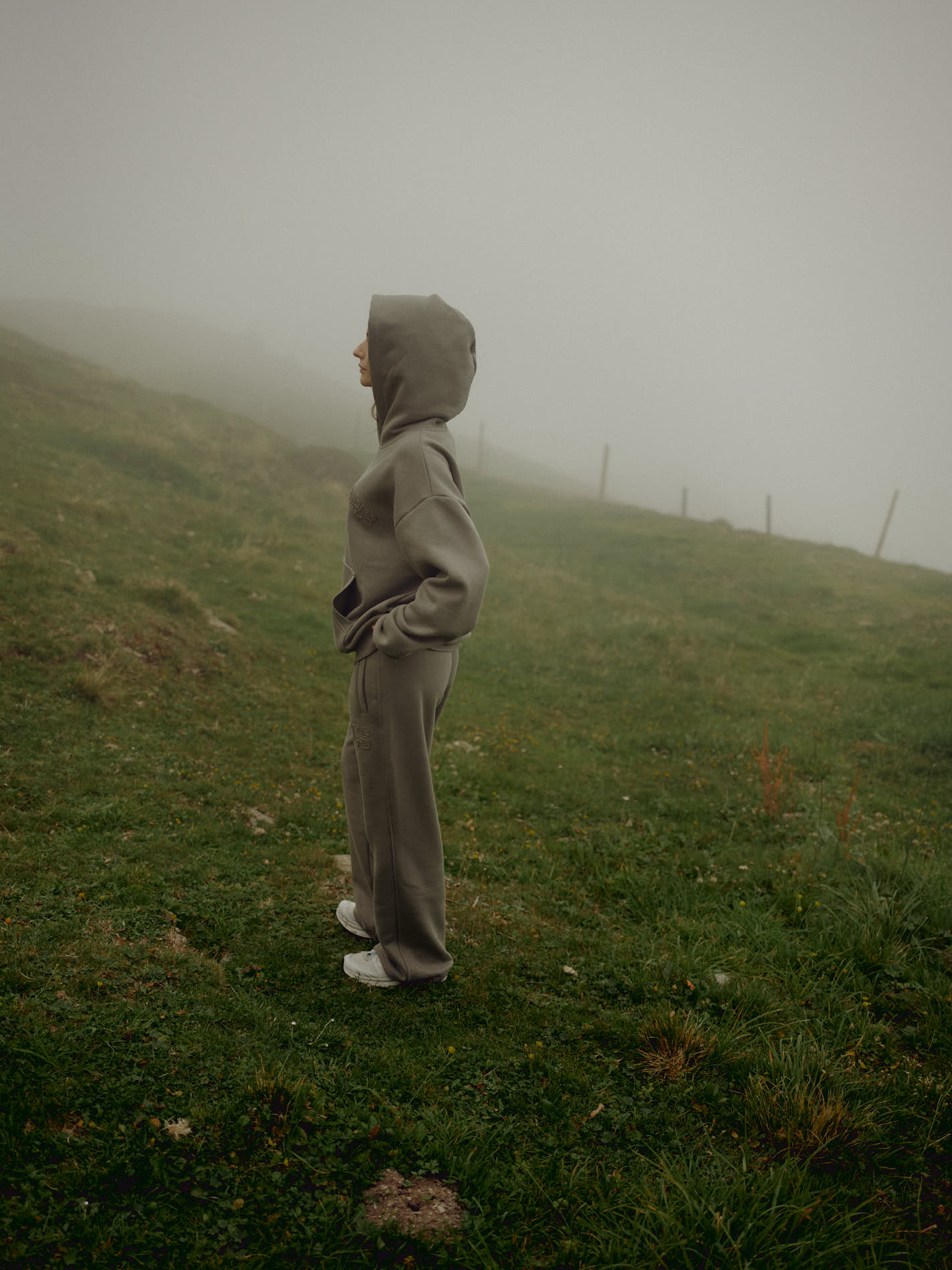Woman wearing a khaki CASPARA CS hoodie with the hood up on a foggy grassy hill.