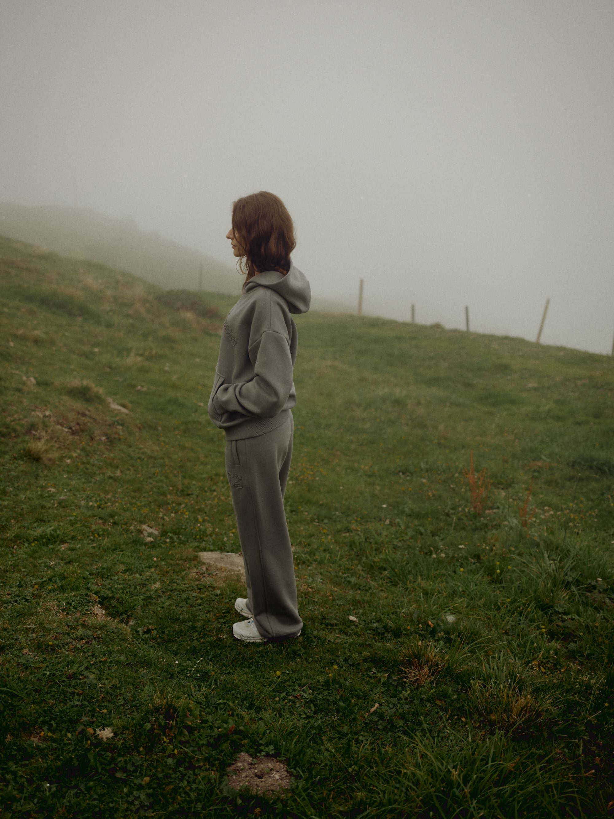 Woman wearing a khaki CASPARA CS hoodie standing on a foggy grassy hillside.