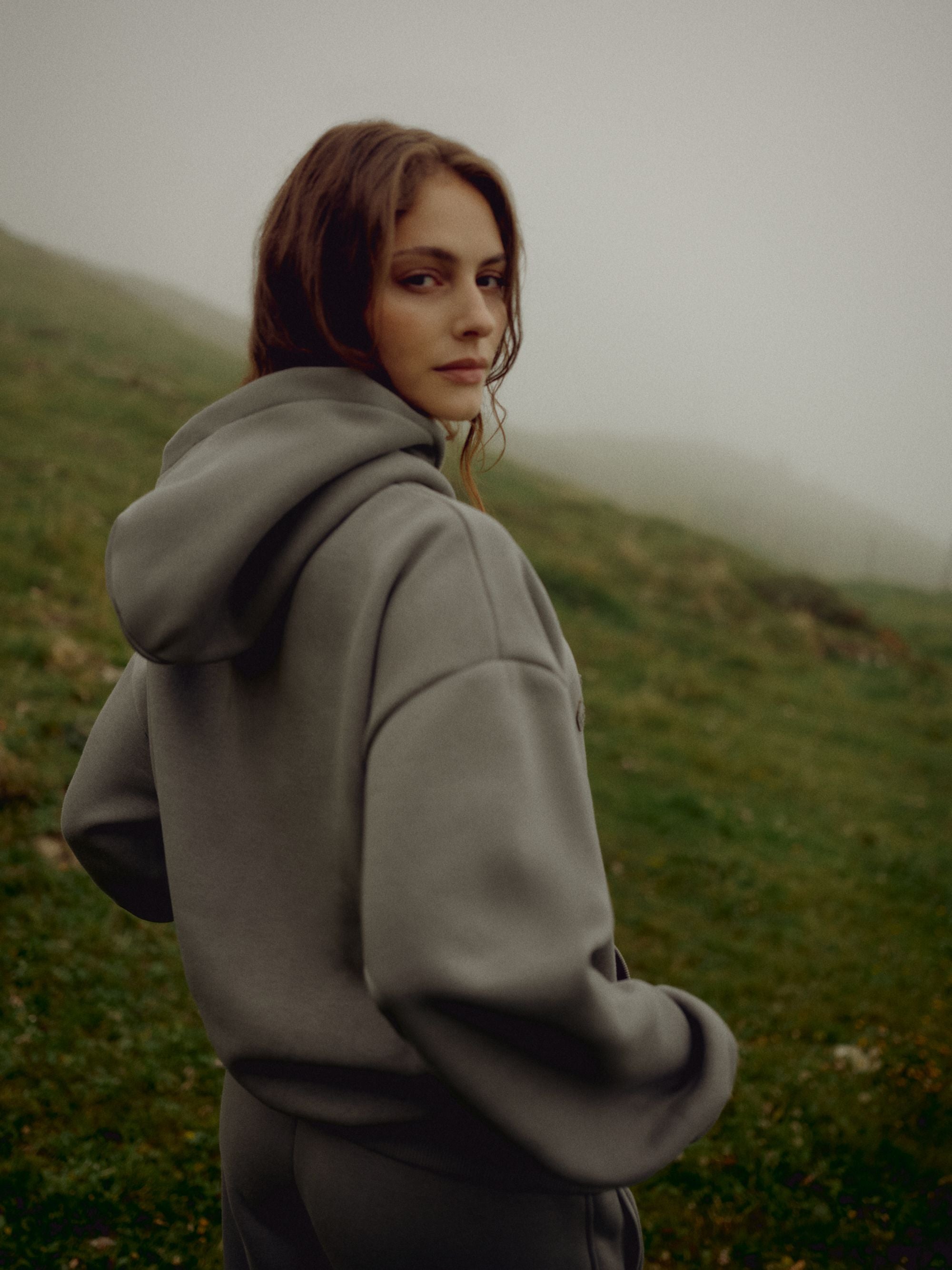 Woman wearing a khaki CASPARA CS hoodie standing on a misty grassy hillside.
