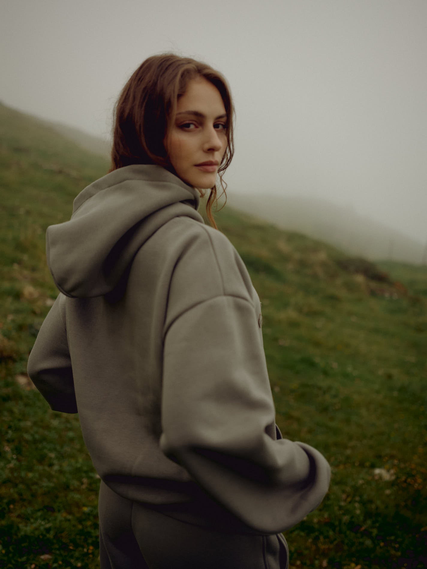 Woman wearing a khaki CASPARA CS hoodie on a misty, grassy hillside.