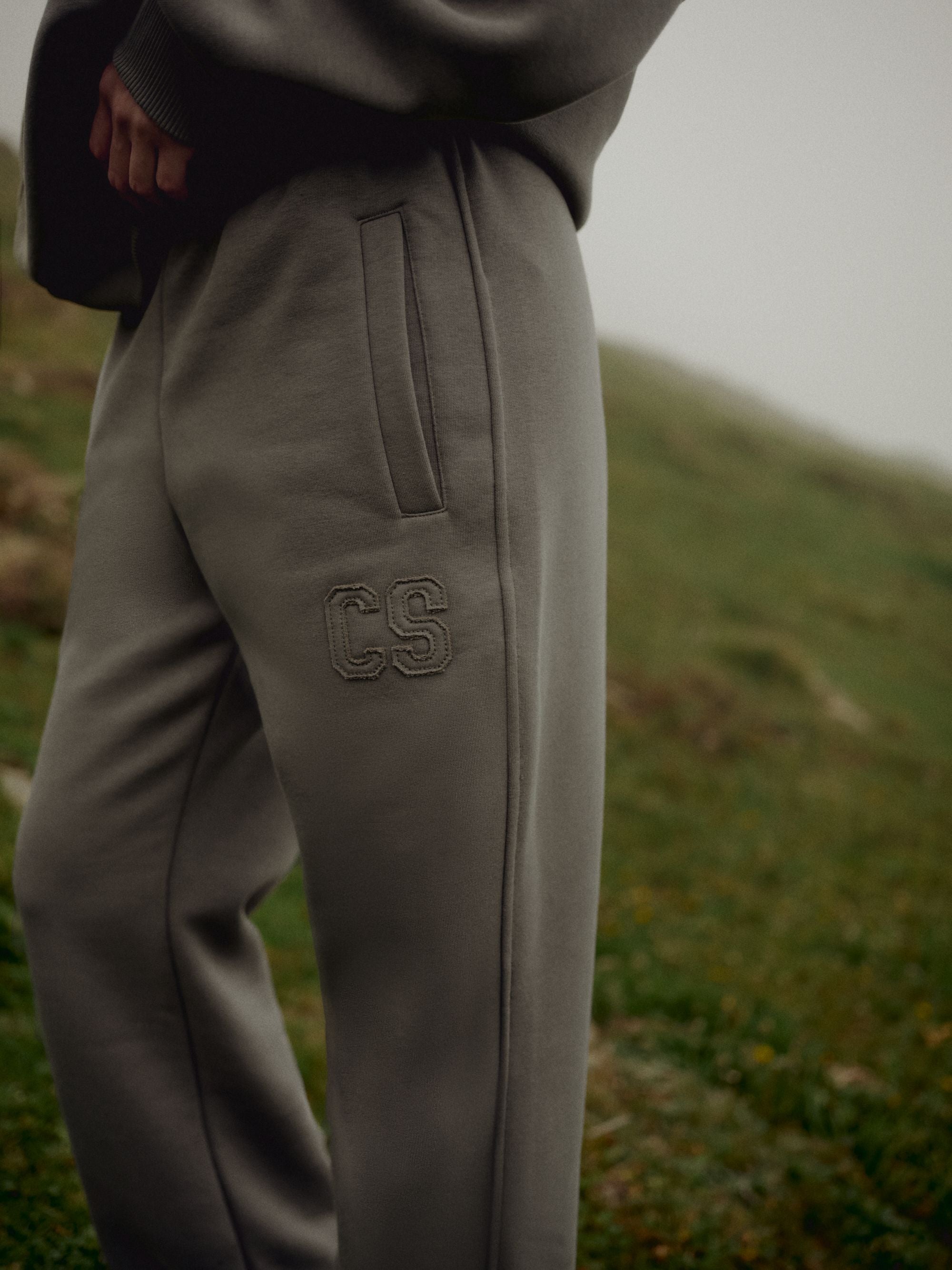 Close-up of khaki CASPARA CS SL sweatpants with embroidered logo on a grassy hillside.