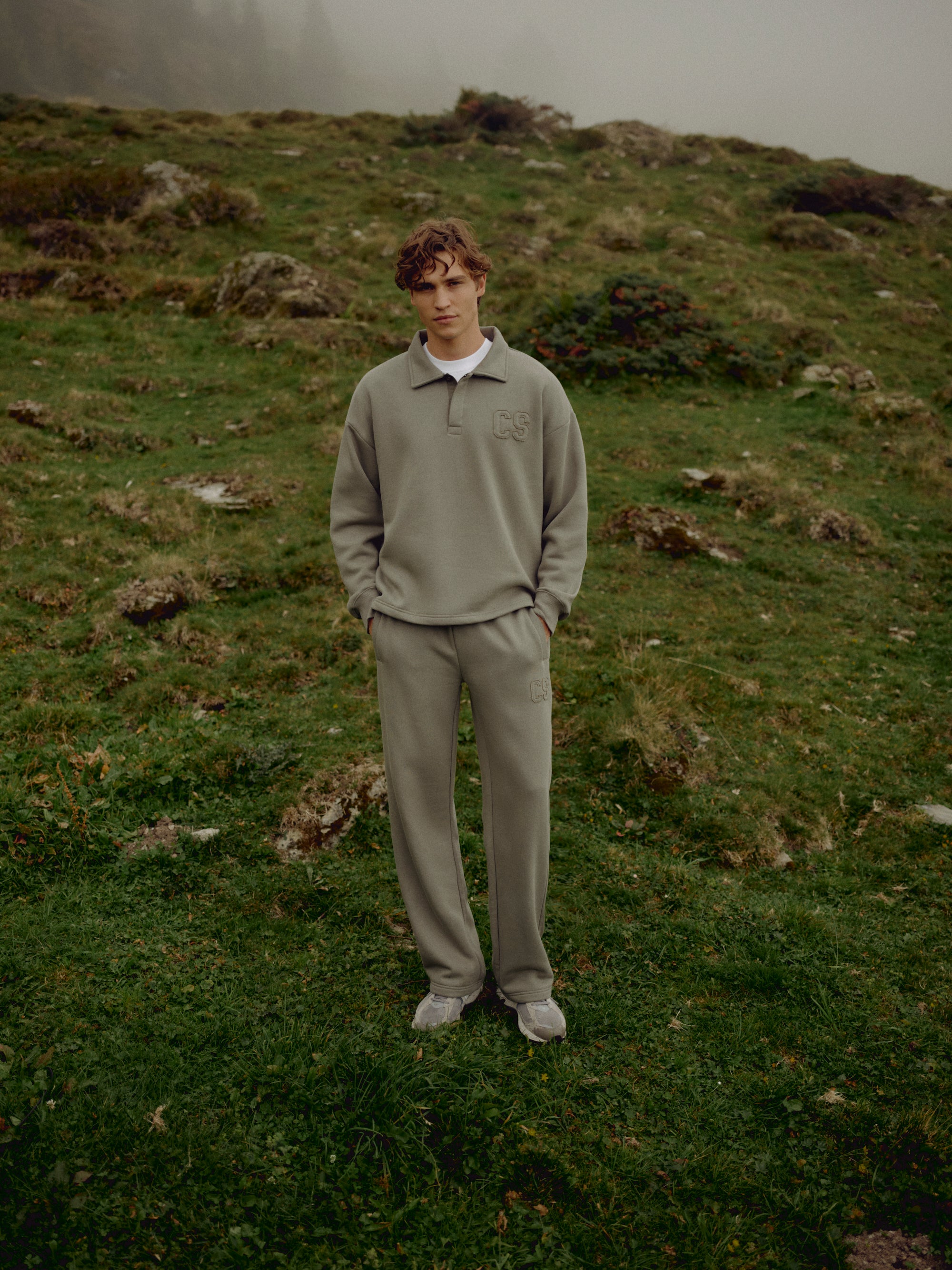 Man wearing a khaki CASPARA CS rugby shirt standing on a grassy hillside.