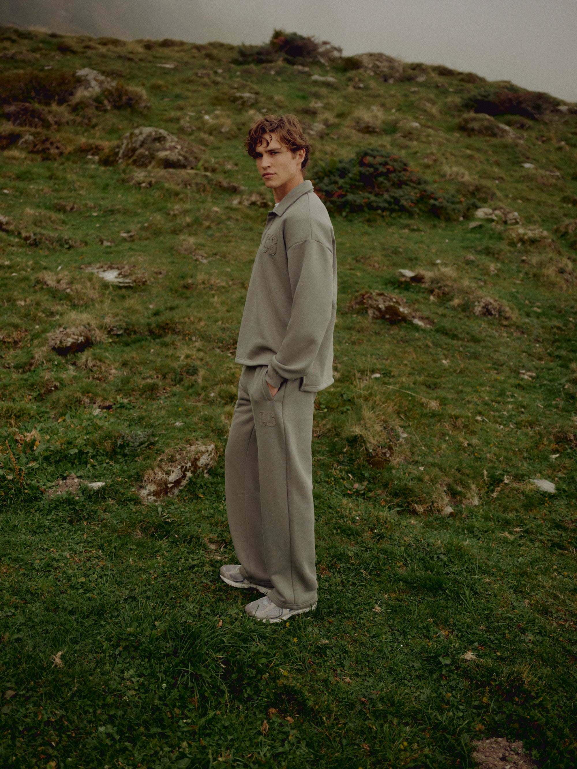 Man wearing a khaki CASPARA CS rugby shirt and sweatpants on a grassy hillside.