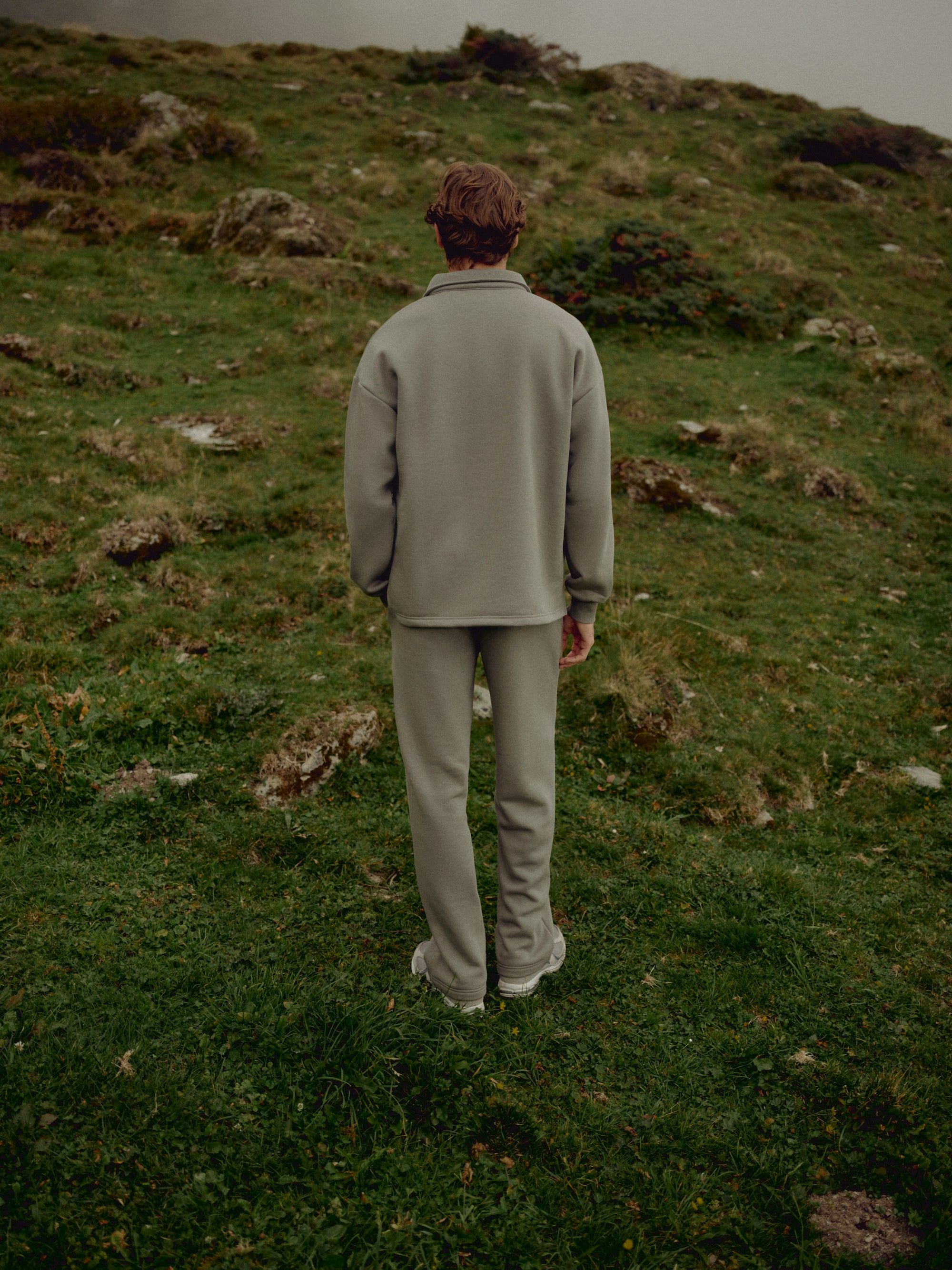 Man wearing khaki CASPARA CS sweatpants on a grassy hillside, viewed from behind.