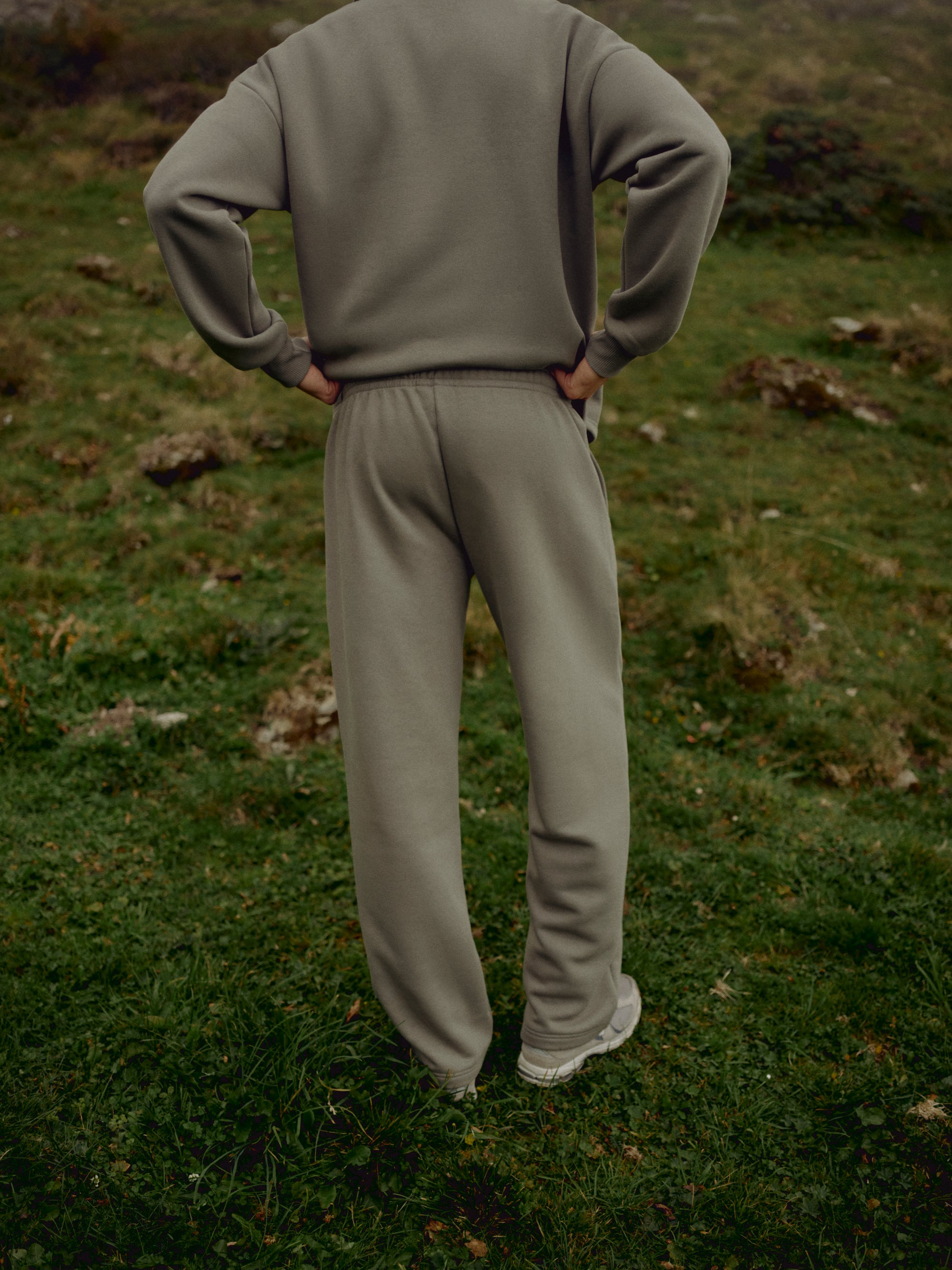 Man wearing khaki CASPARA CS sweatpants standing in a grassy field, viewed from behind.
