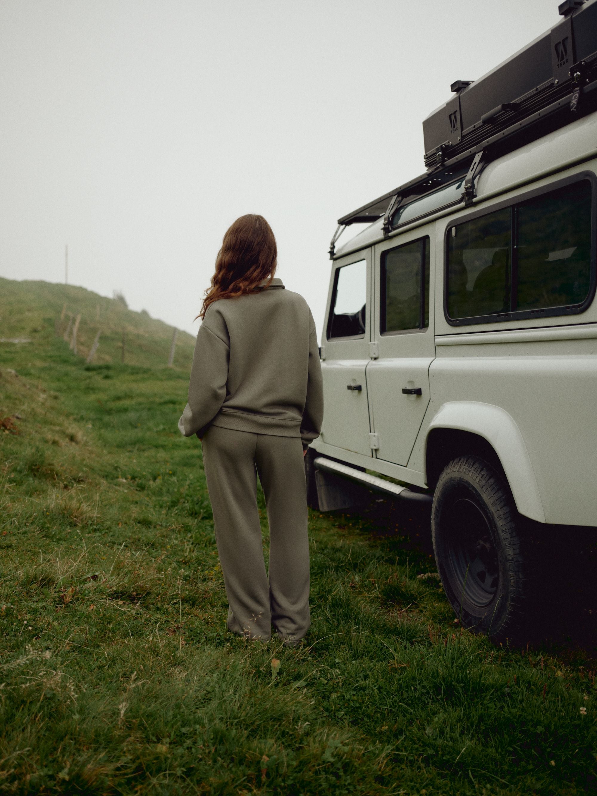 Woman wearing a khaki CASPARA CS rugby shirt and sweatpants beside an off-road vehicle on a misty hill.