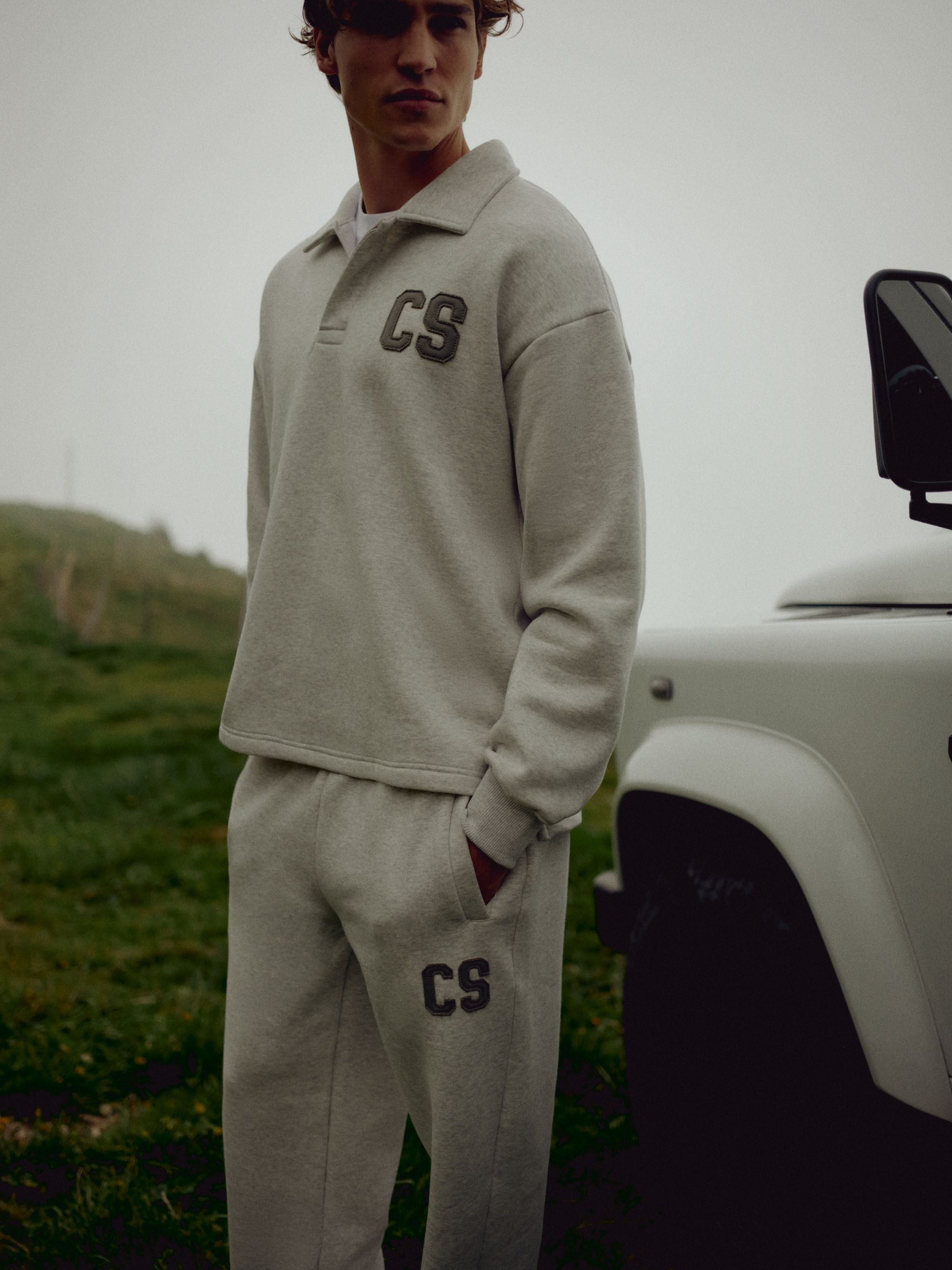 Man wearing a grey melange CASPARA CS rugby shirt and sweatpants outdoors next to a car.