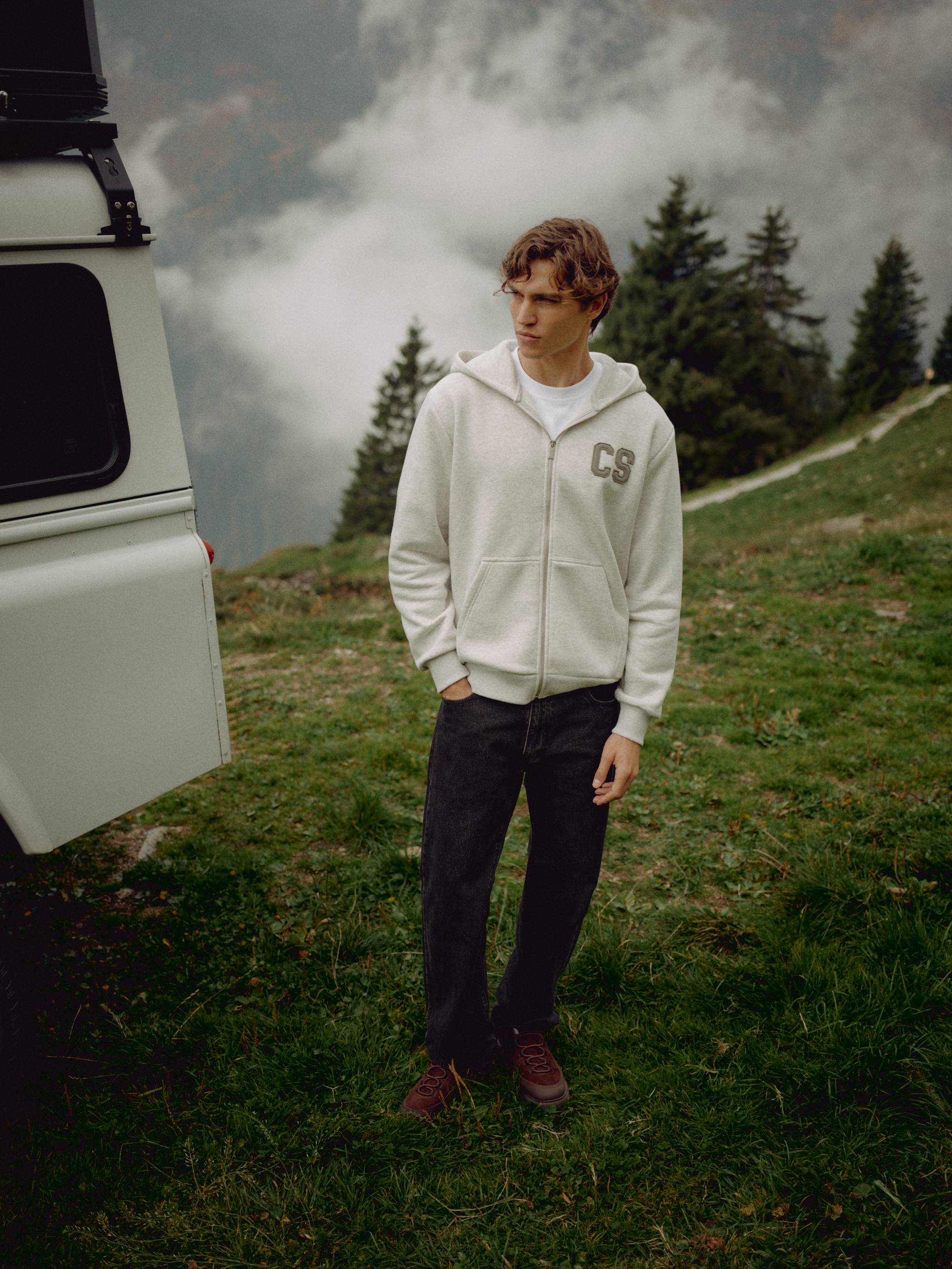 Man wearing a grey melange CASPARA CS zip hoodie standing beside a vehicle on a mountain hillside.
