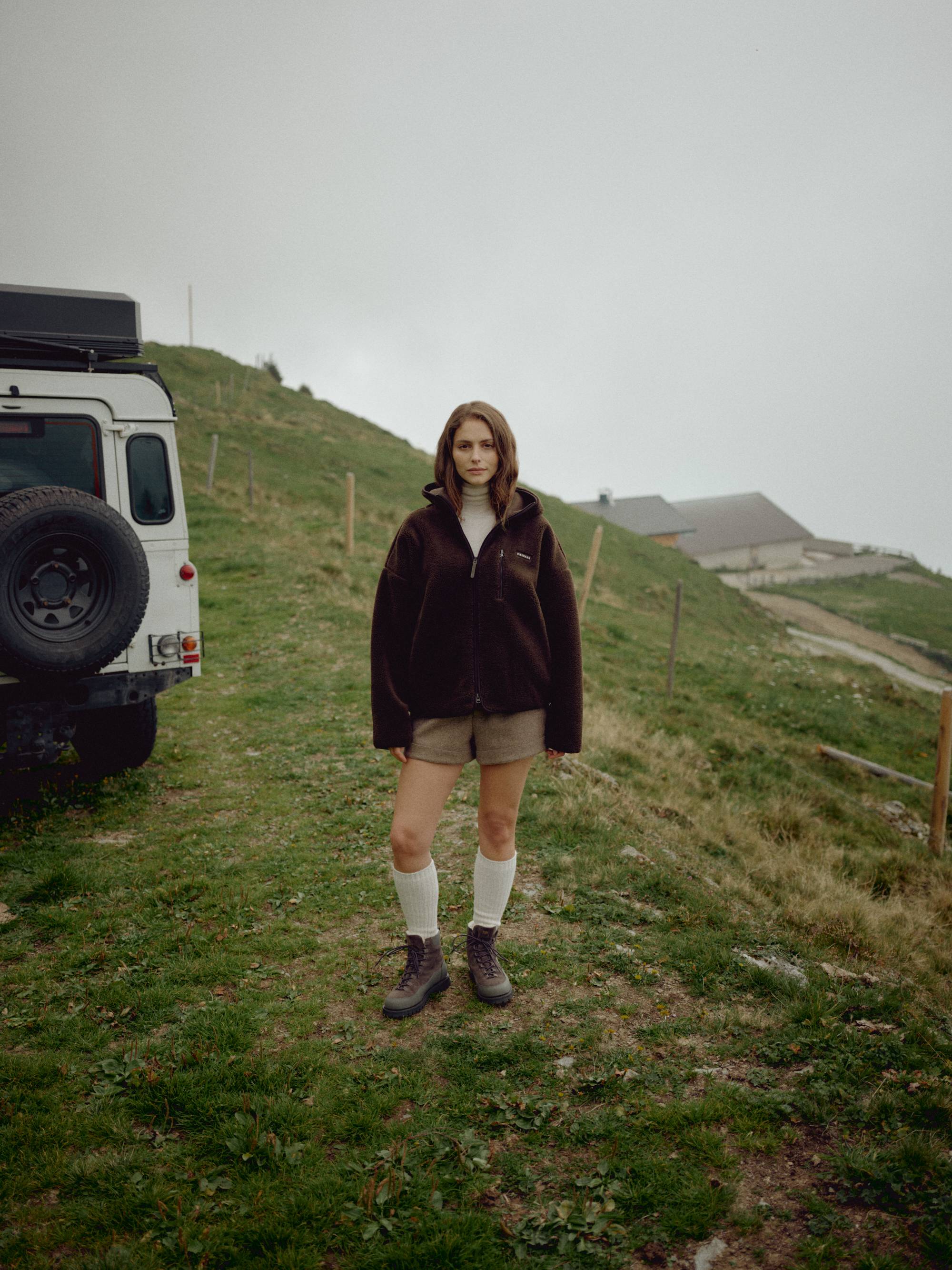 Woman wearing an espresso CASPARA Cloudfleece hooded jacket on a grassy hillside next to a vehicle.