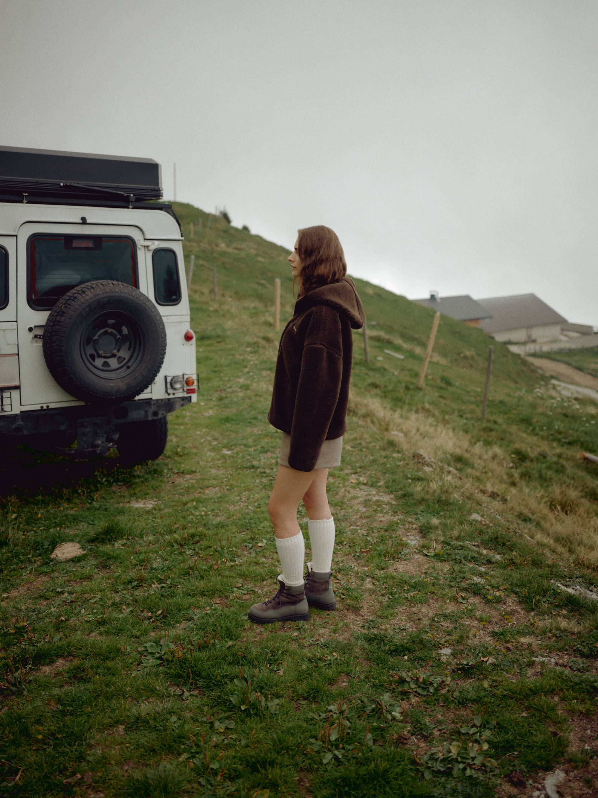 Woman wearing an espresso CASPARA Cloudfleece hooded jacket next to an off-road vehicle.