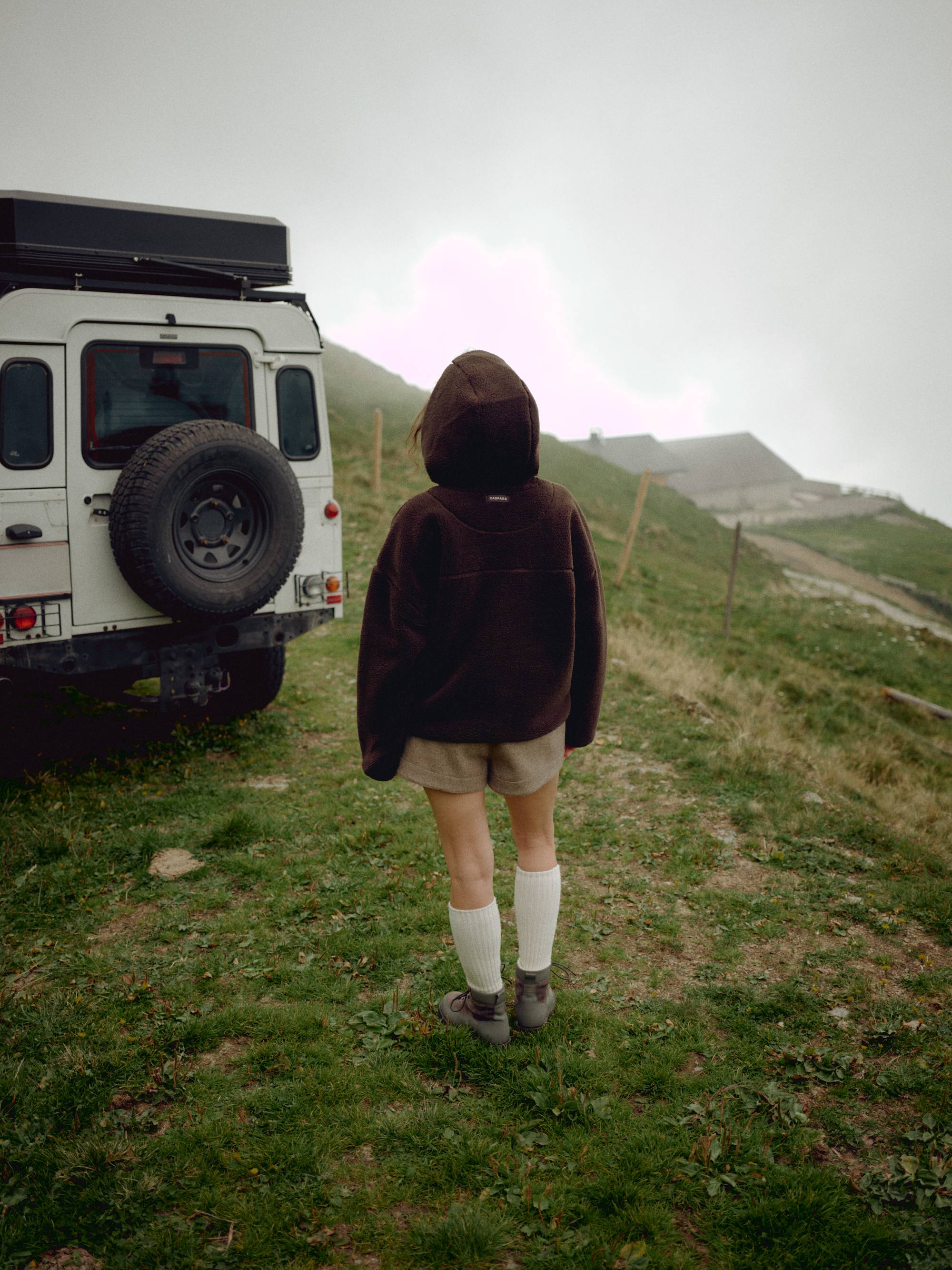 Woman wearing an espresso CASPARA Cloudfleece hooded jacket next to an off-road vehicle in the mountains.