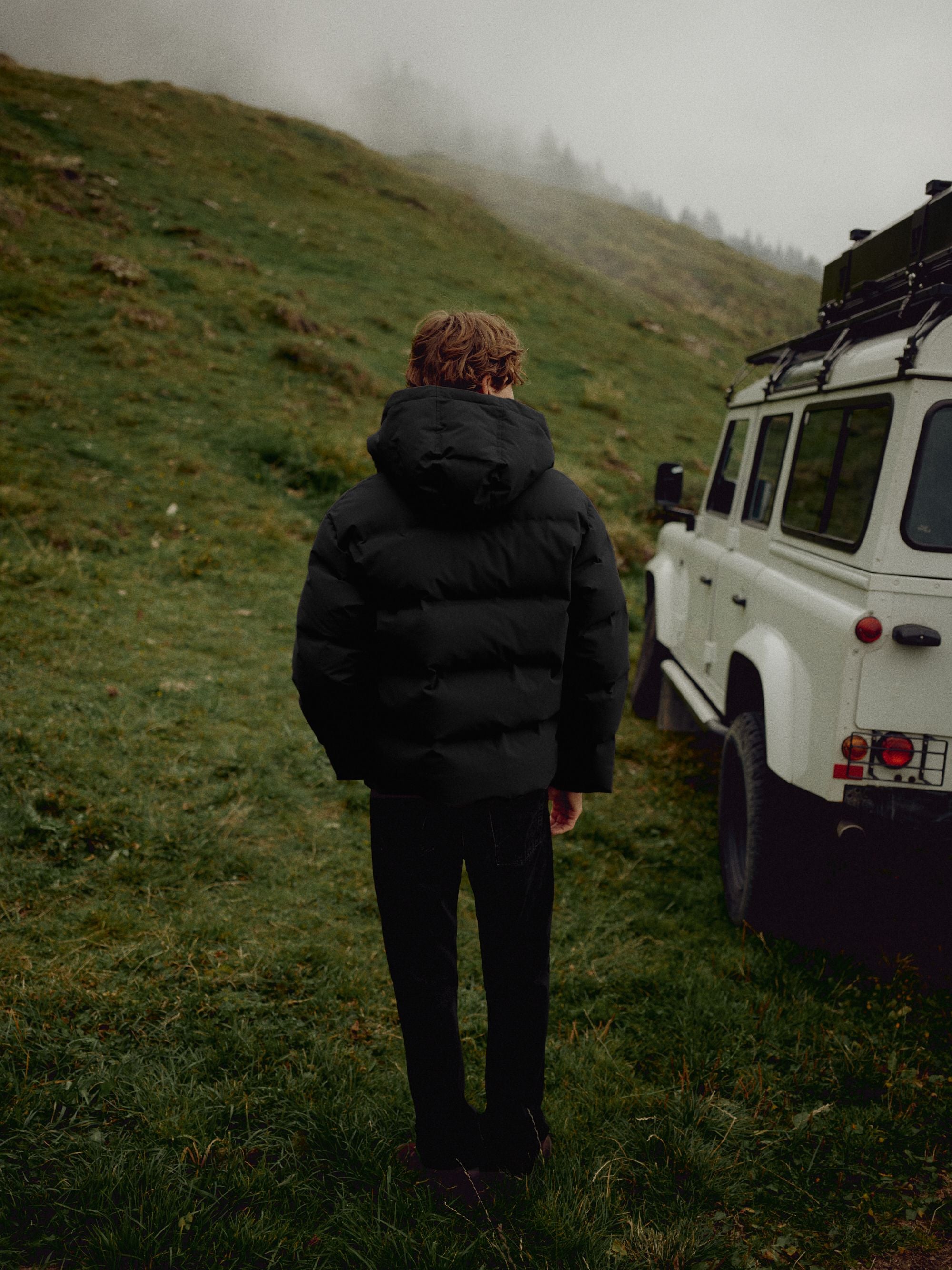 Back view of a man wearing a black CASPARA Boreal puffer jacket outdoors next to an off-road vehicle.