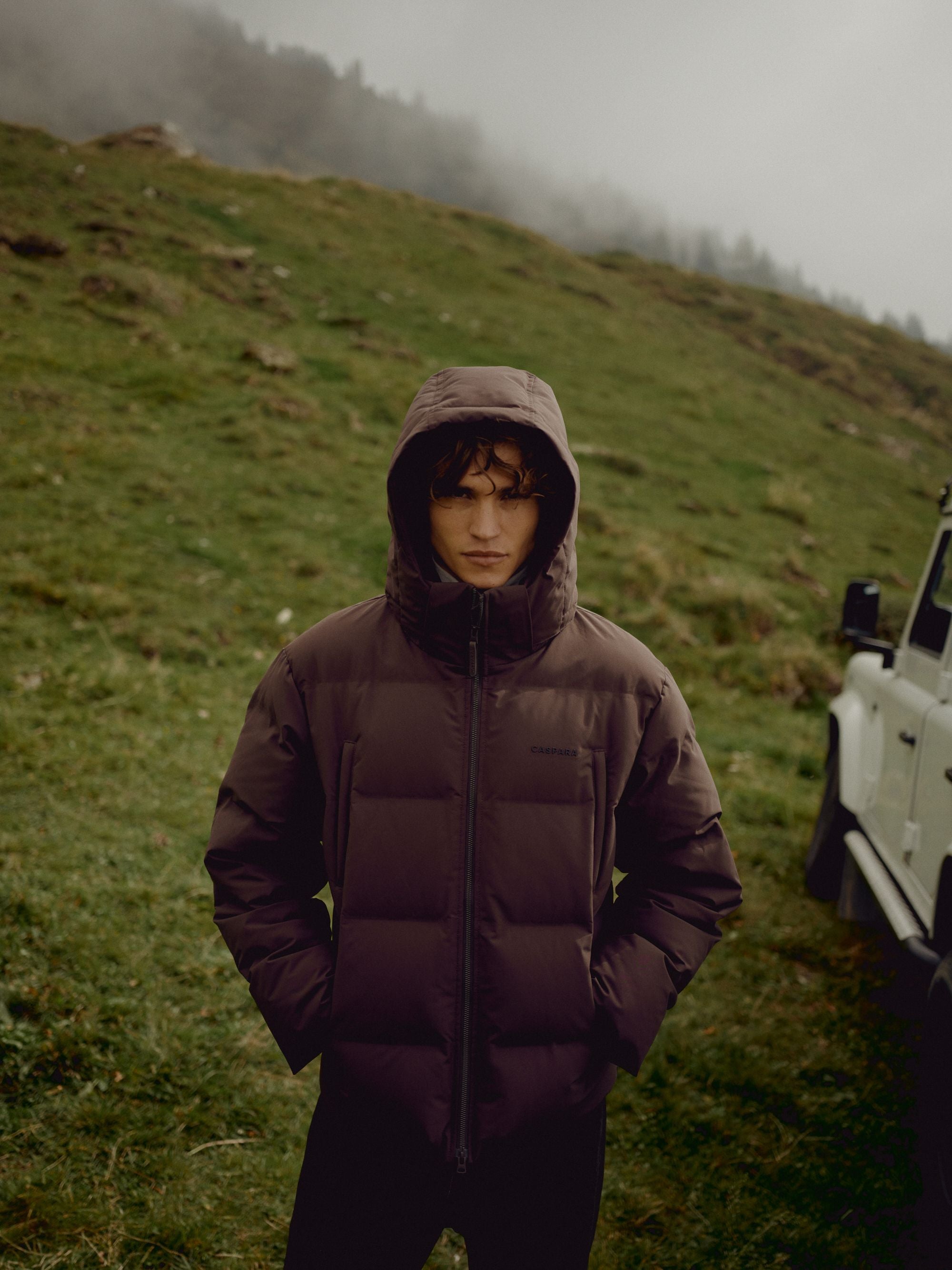 Man wearing a cocoa bean CASPARA Boreal puffer jacket outdoors on a hillside.