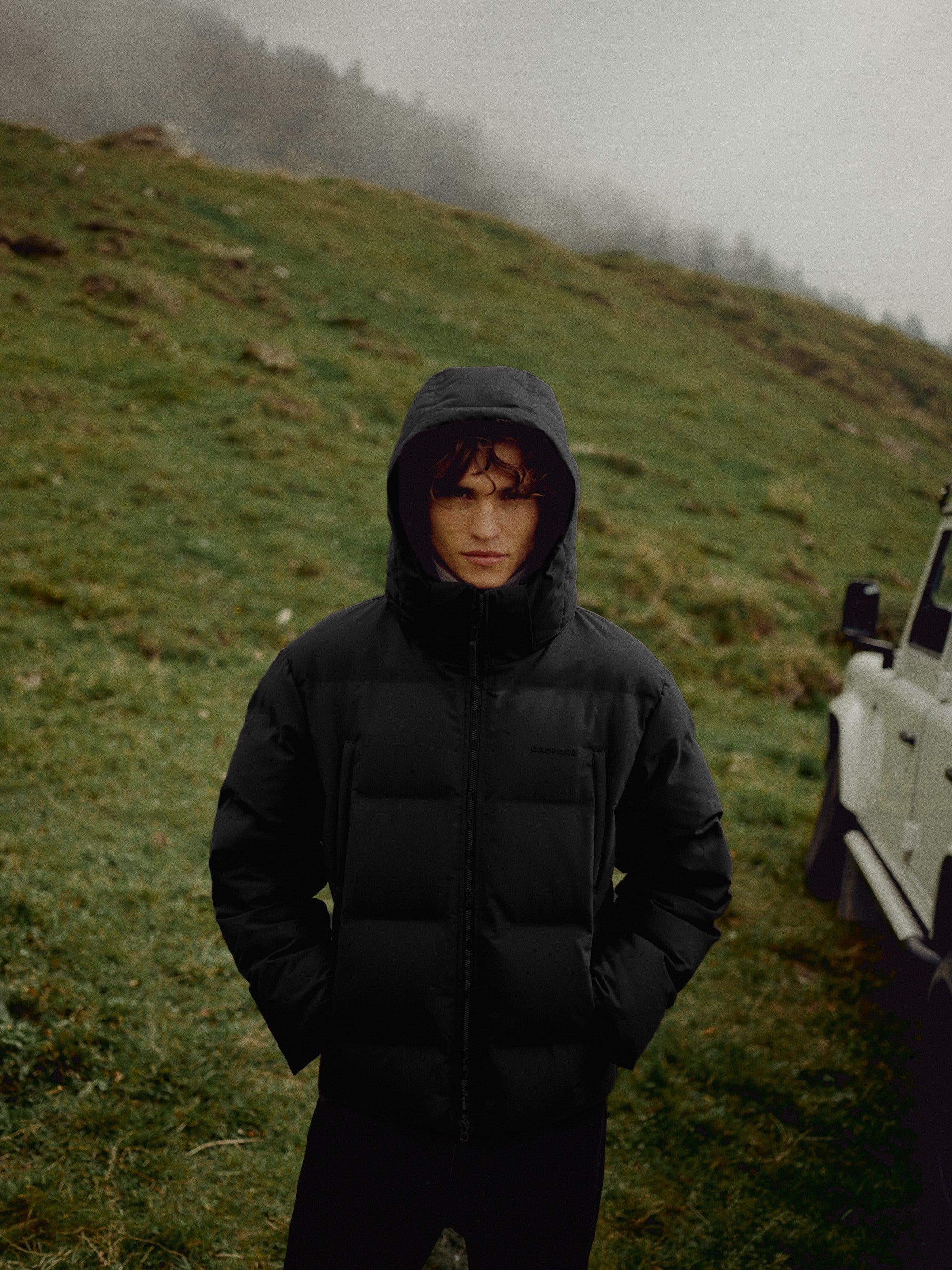 Man wearing a black CASPARA Boreal puffer jacket outdoors on a foggy hillside.