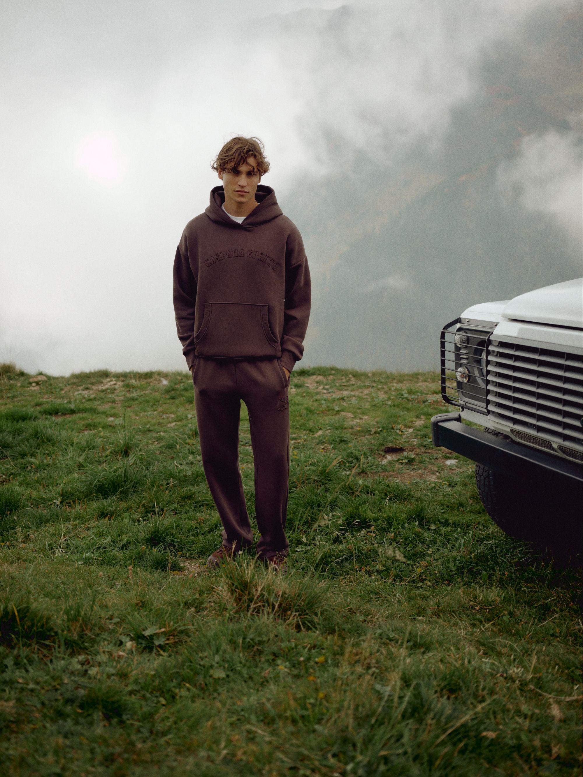 Man wearing an espresso CASPARA CS hoodie and sweatpants standing in a mountain landscape.