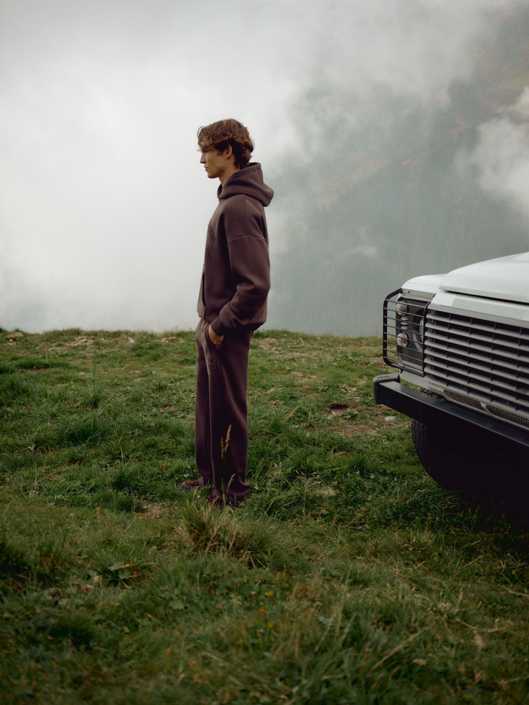 Man wearing an espresso CASPARA CS hoodie and sweatpants on a misty mountain hillside.