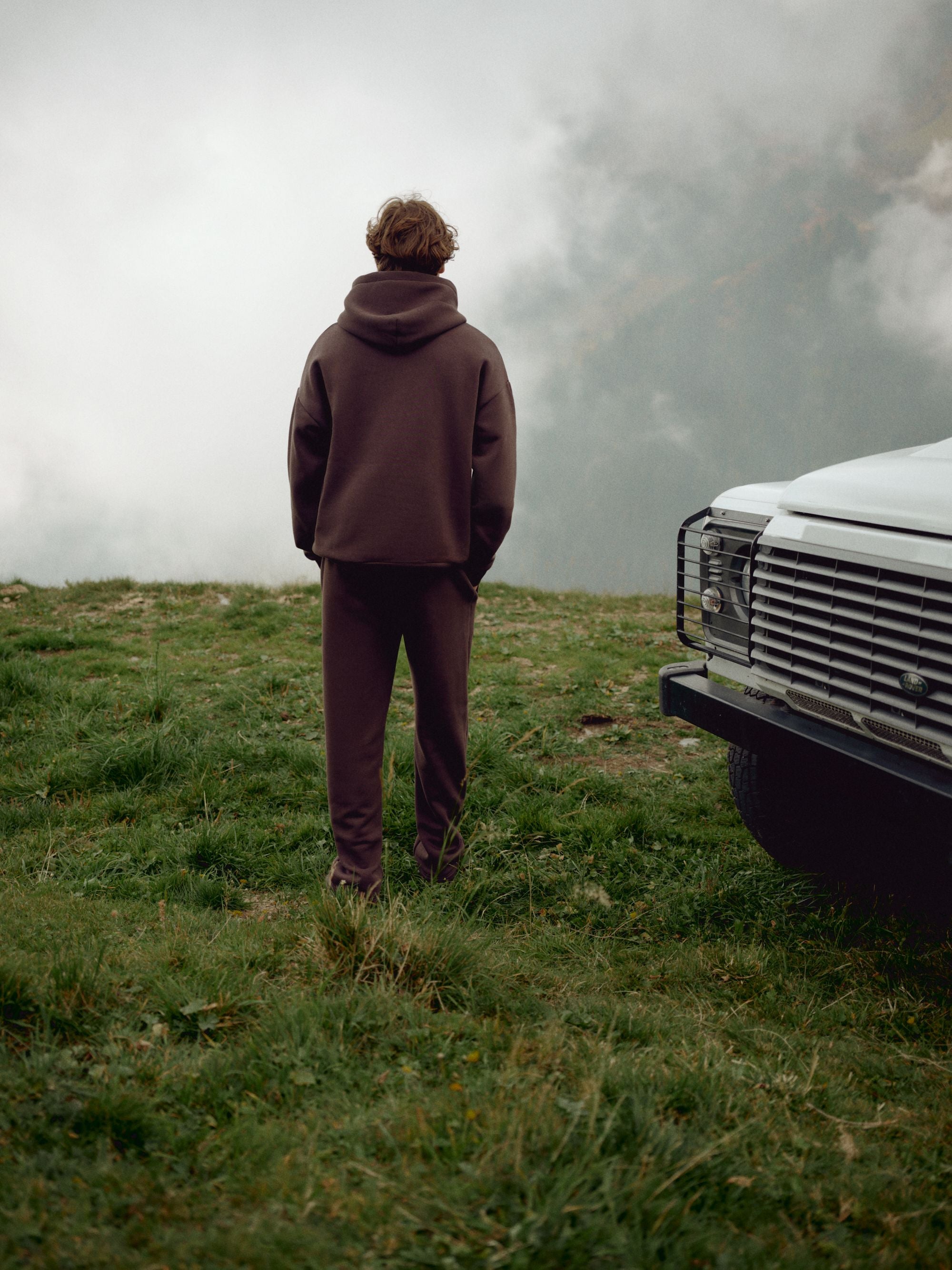 Man wearing an espresso CASPARA CS hoodie and sweatpants next to a vehicle on a misty hillside.