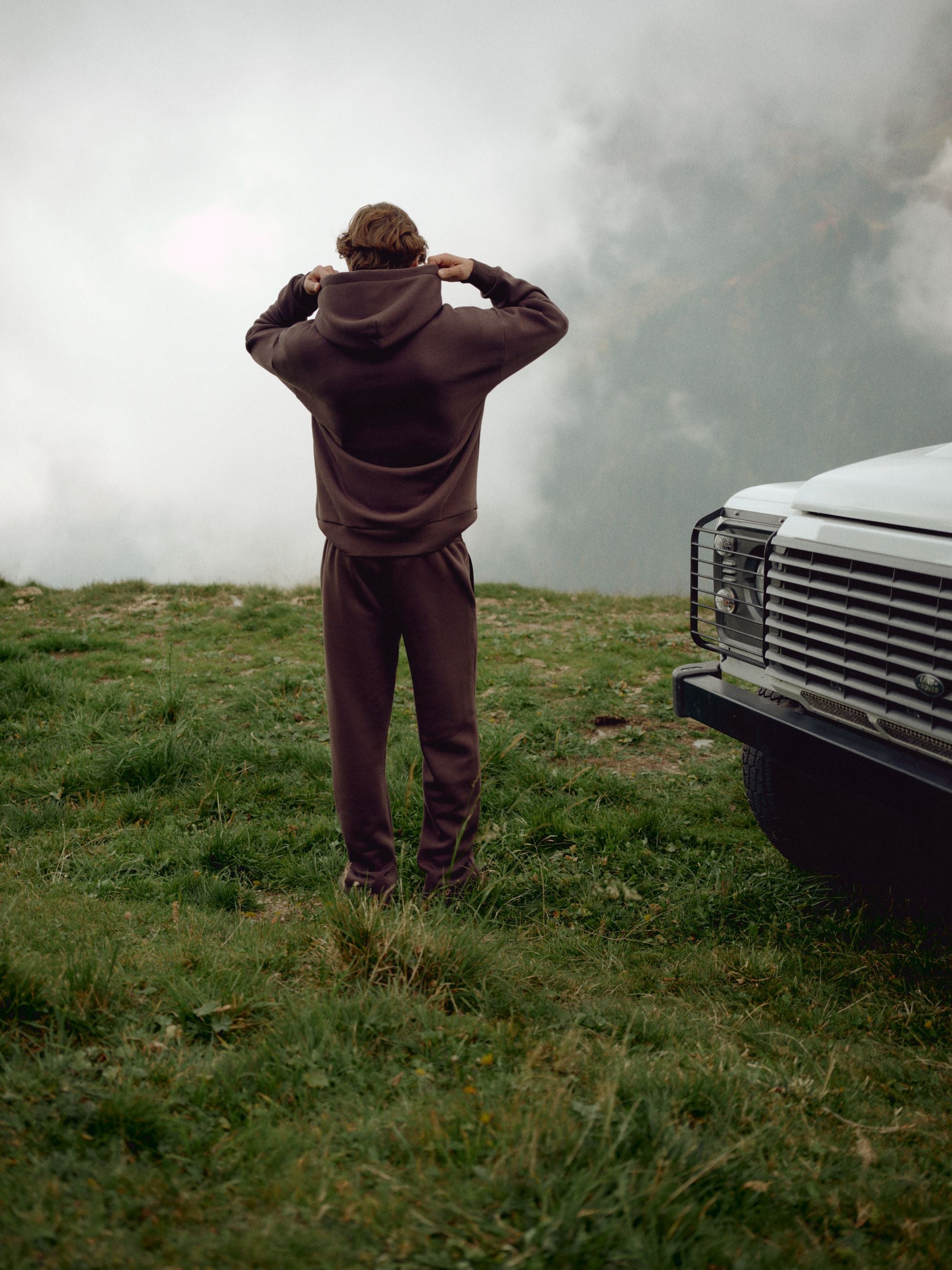 Man wearing an espresso CASPARA CS hoodie and sweatpants on a misty hillside by an off-road vehicle.