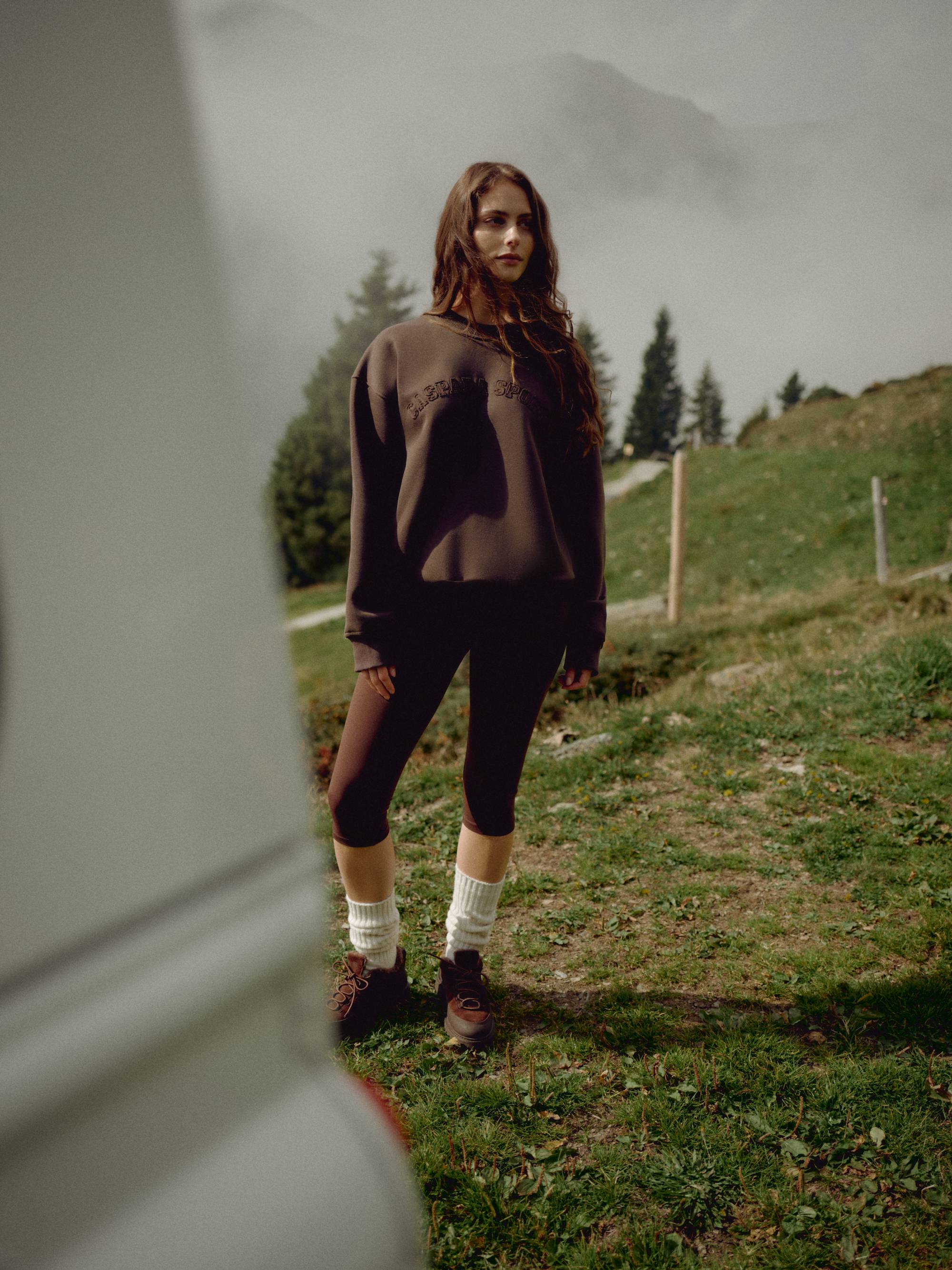 Woman wearing an espresso CASPARA CS crewneck sweatshirt standing on a grassy hillside.