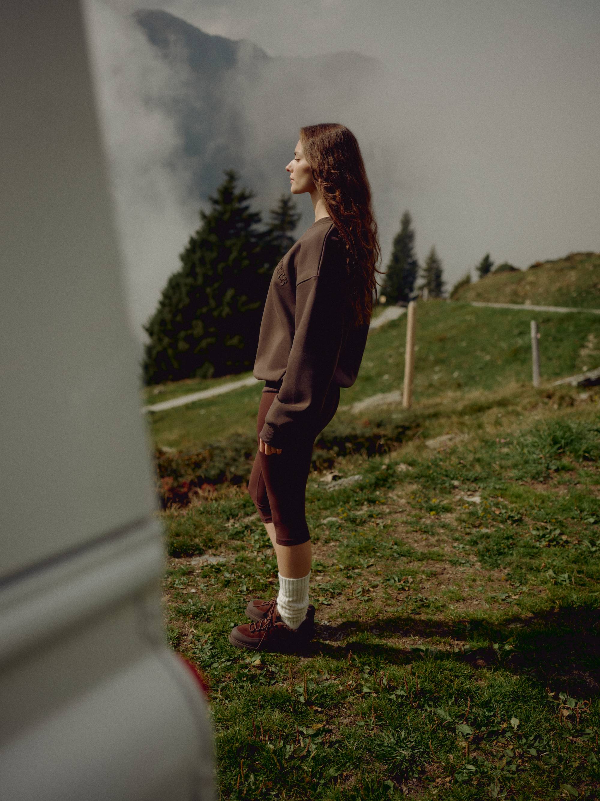 Woman wearing an espresso CASPARA CS crewneck sweatshirt standing in a misty mountain landscape.