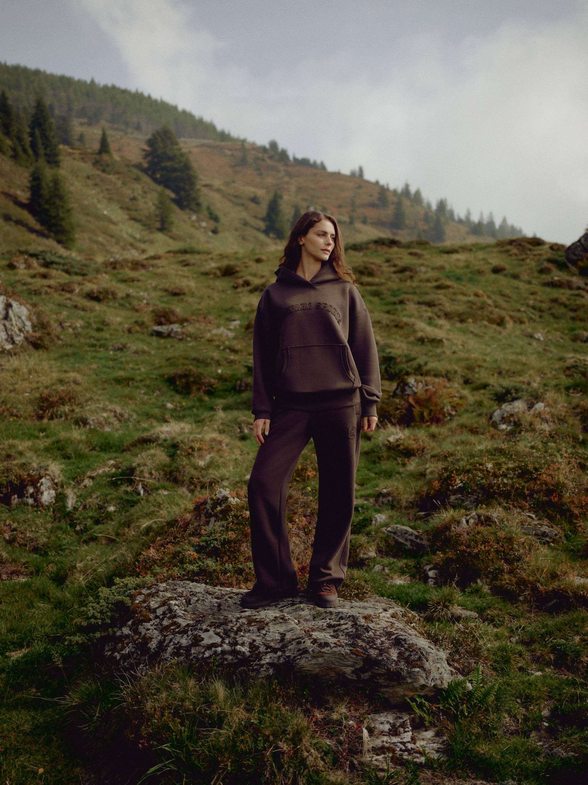 Woman wearing an espresso CASPARA CS hoodie and sweatpants standing on a rocky hillside.