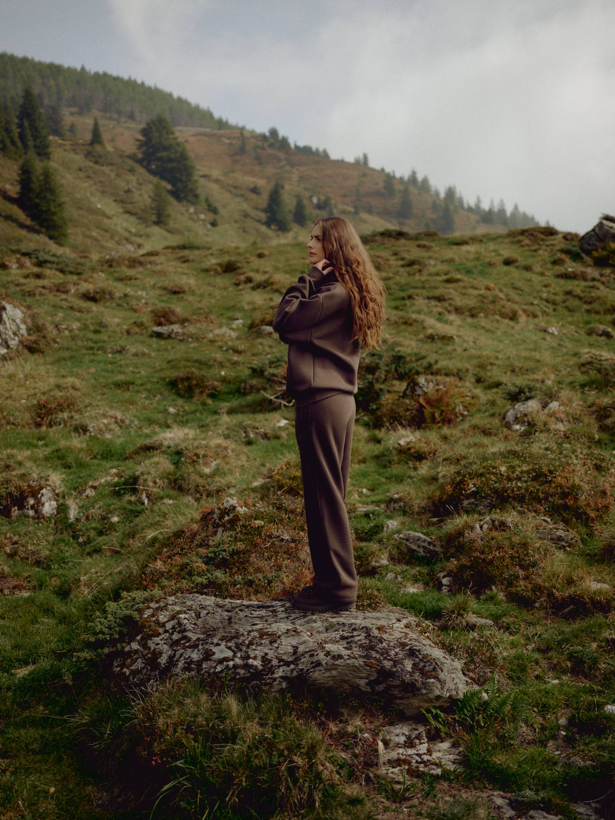 Woman wearing an espresso CASPARA CS hoodie and sweatpants standing on a hillside.