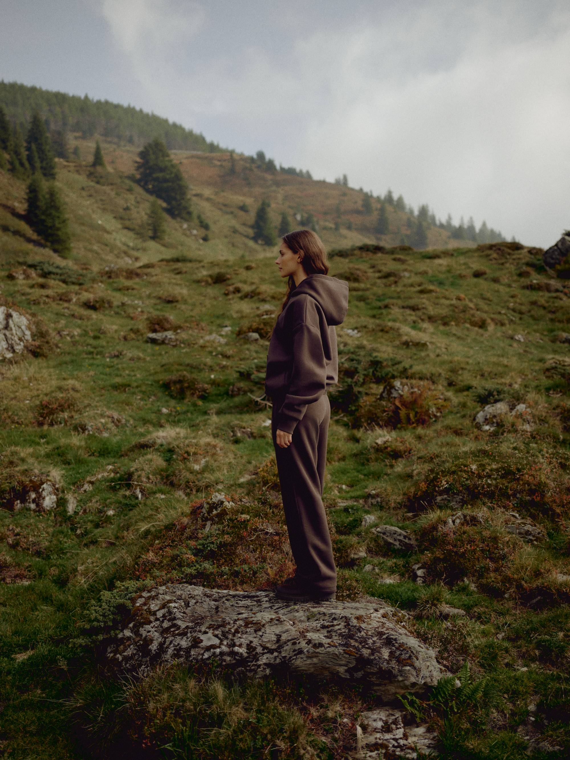 Woman wearing an espresso CASPARA CS hoodie and sweatpants on a grassy hillside.