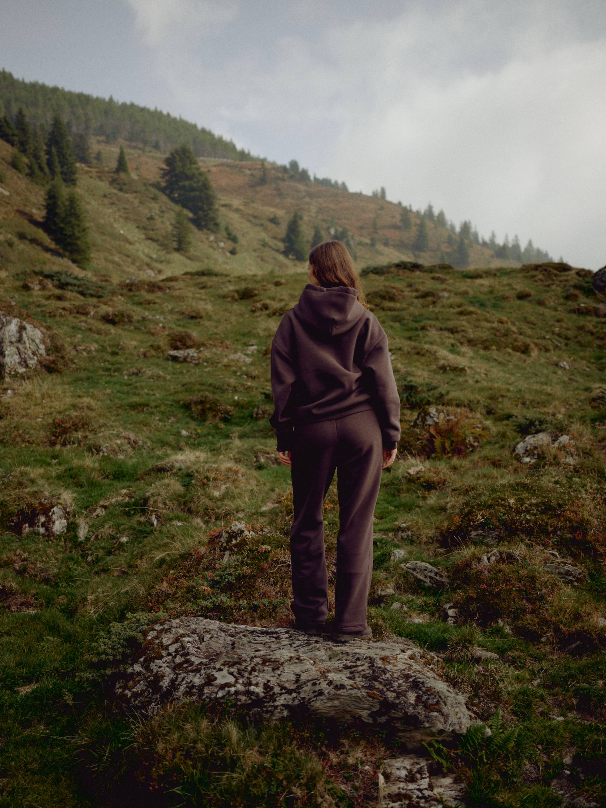 Woman wearing an espresso CASPARA CS hoodie and sweatpants on a grassy hillside.