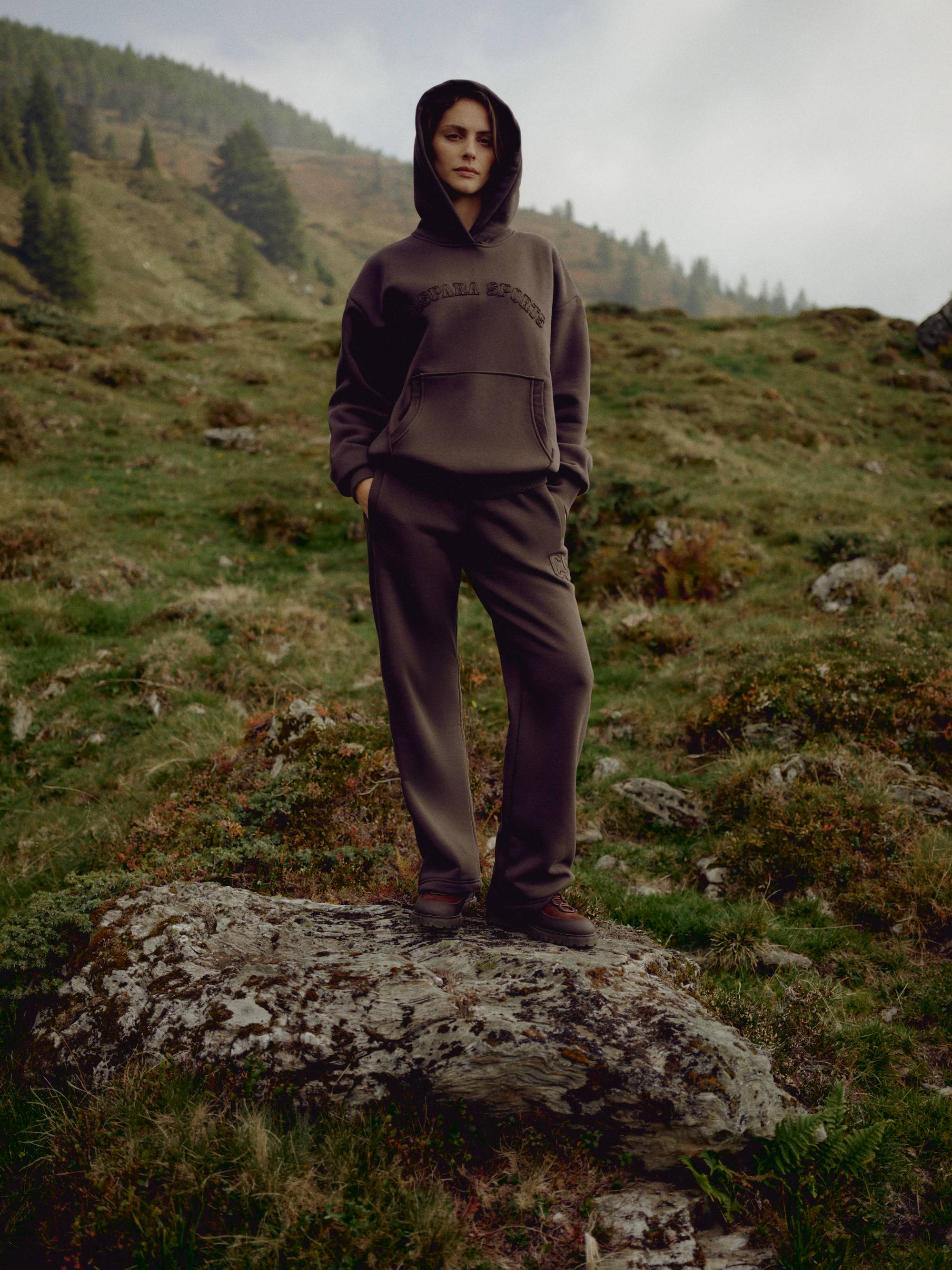 Woman wearing an espresso CASPARA CS hoodie and matching sweatpants on a rocky hillside.