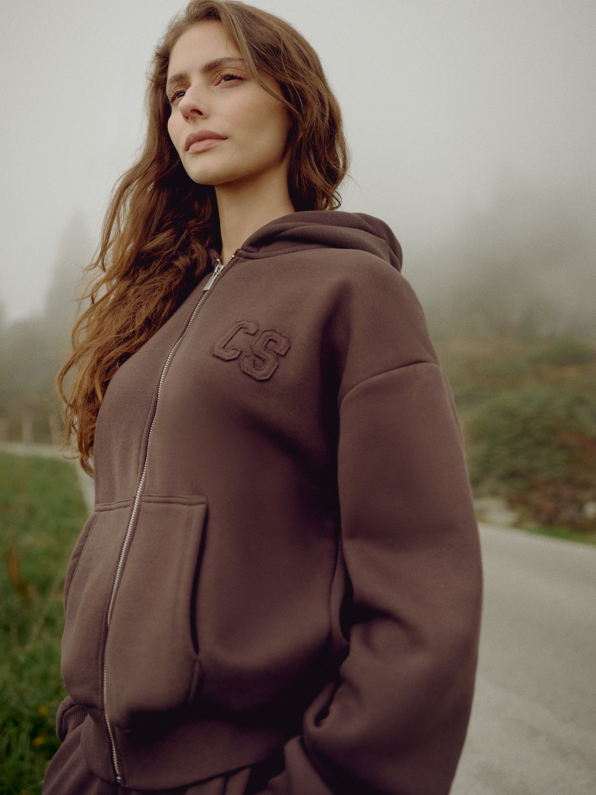 Woman wearing an espresso CASPARA CS zip hoodie on a misty roadside.