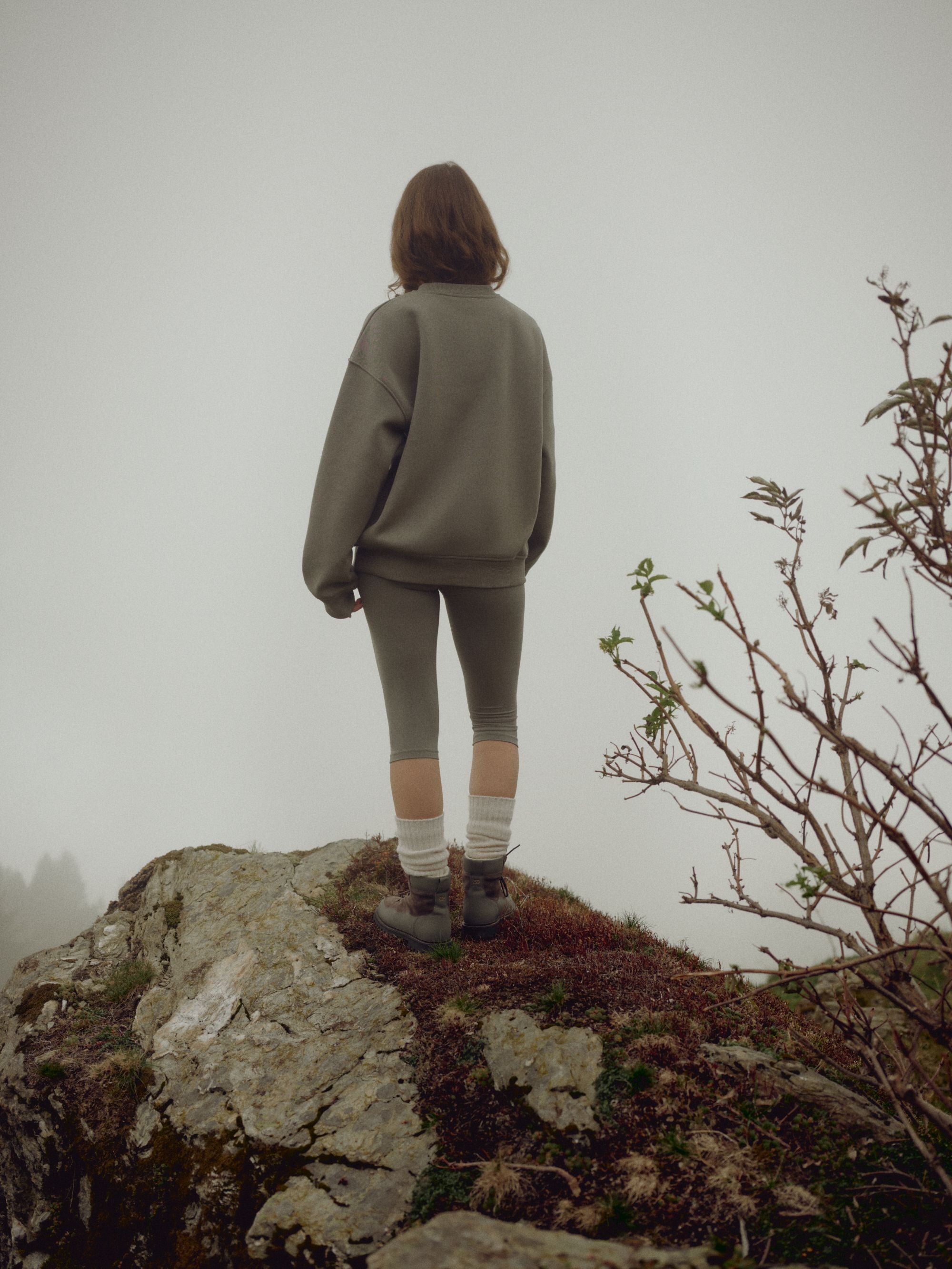 Woman wearing a khaki CASPARA CS crewneck sweatshirt standing on a misty rocky hillside.