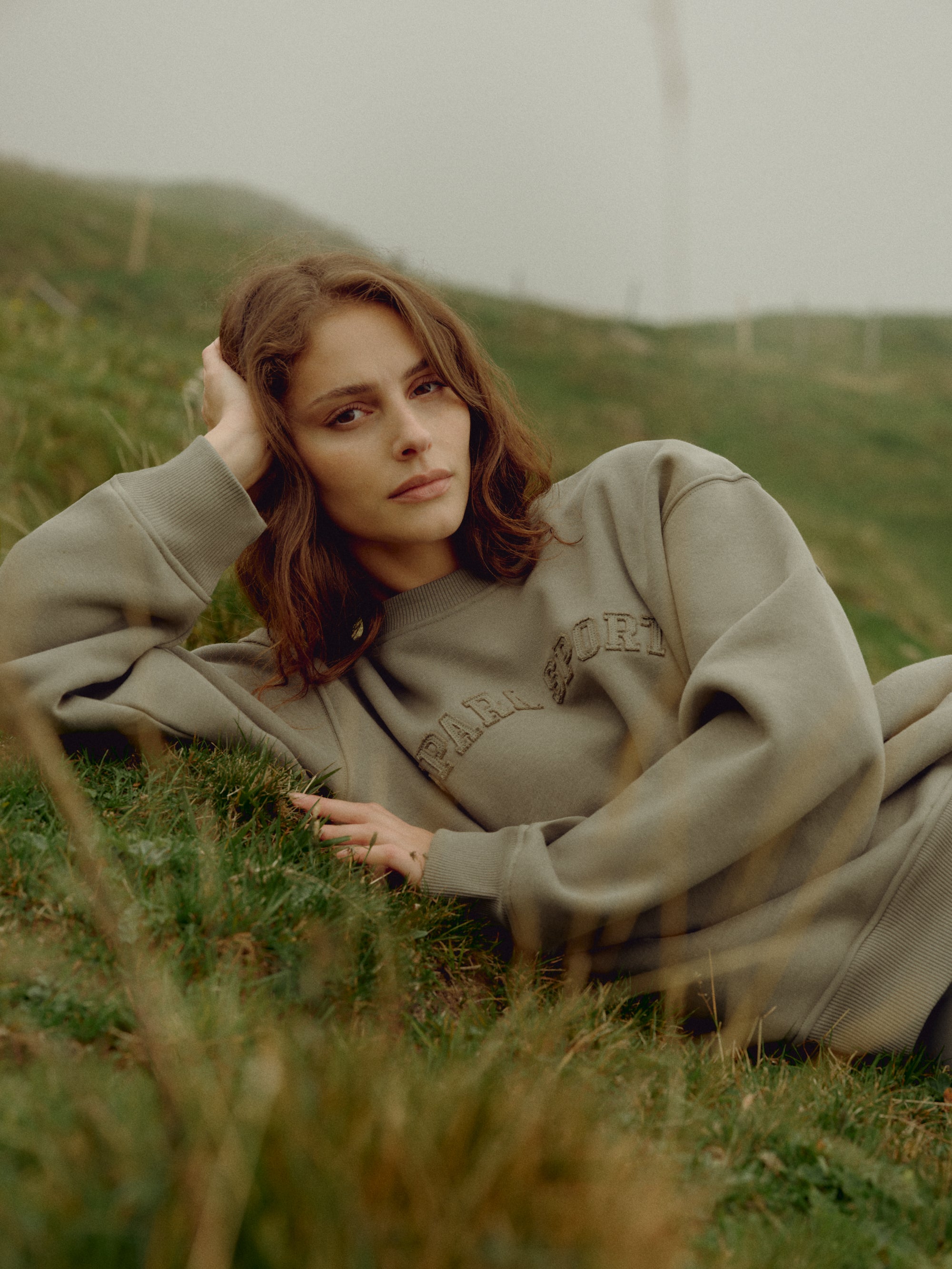 Woman wearing a khaki CASPARA CS crewneck sweatshirt lying on a grassy hillside.