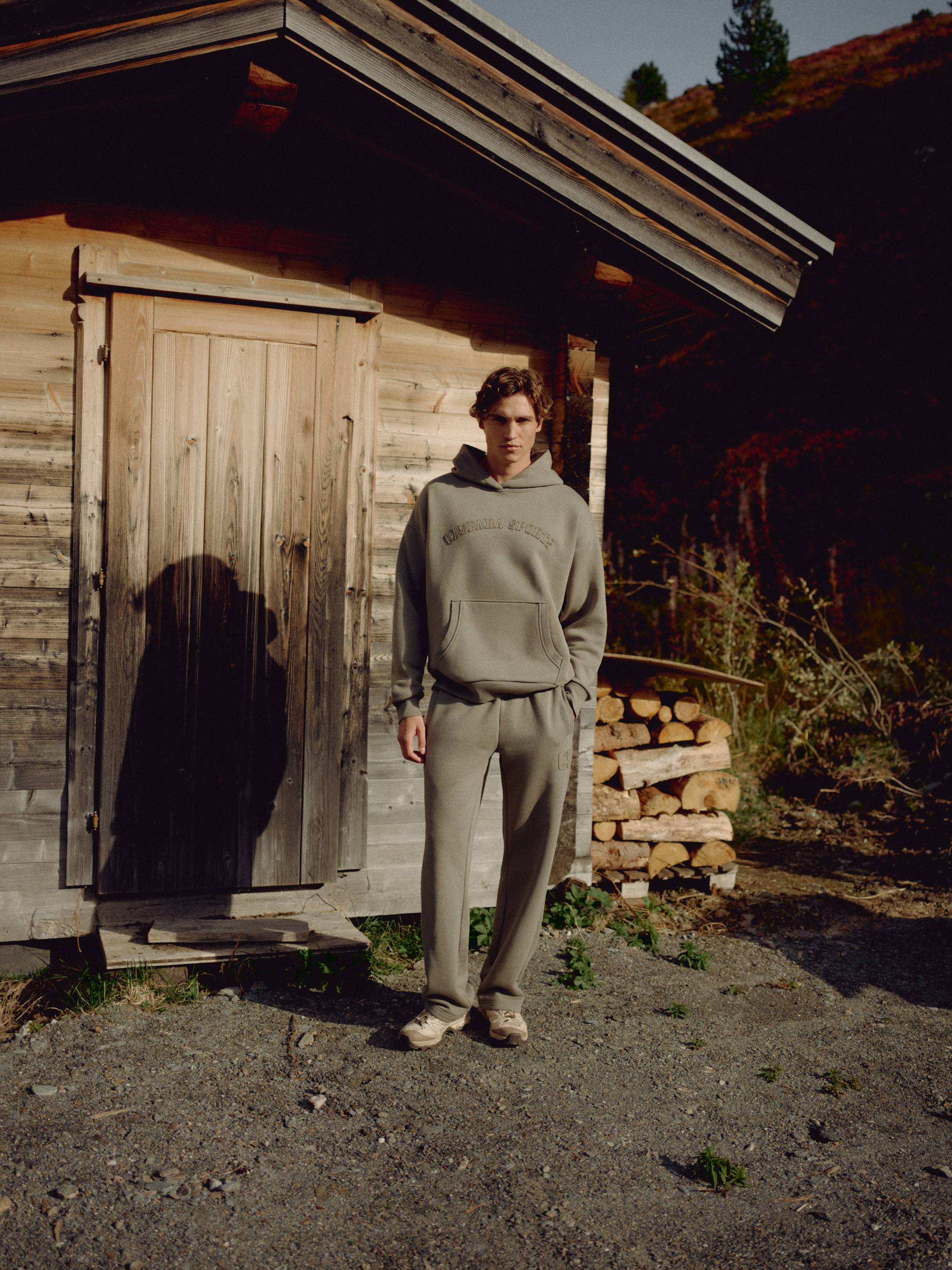 Man wearing a khaki CASPARA CS hoodie standing outside a rustic wooden cabin.