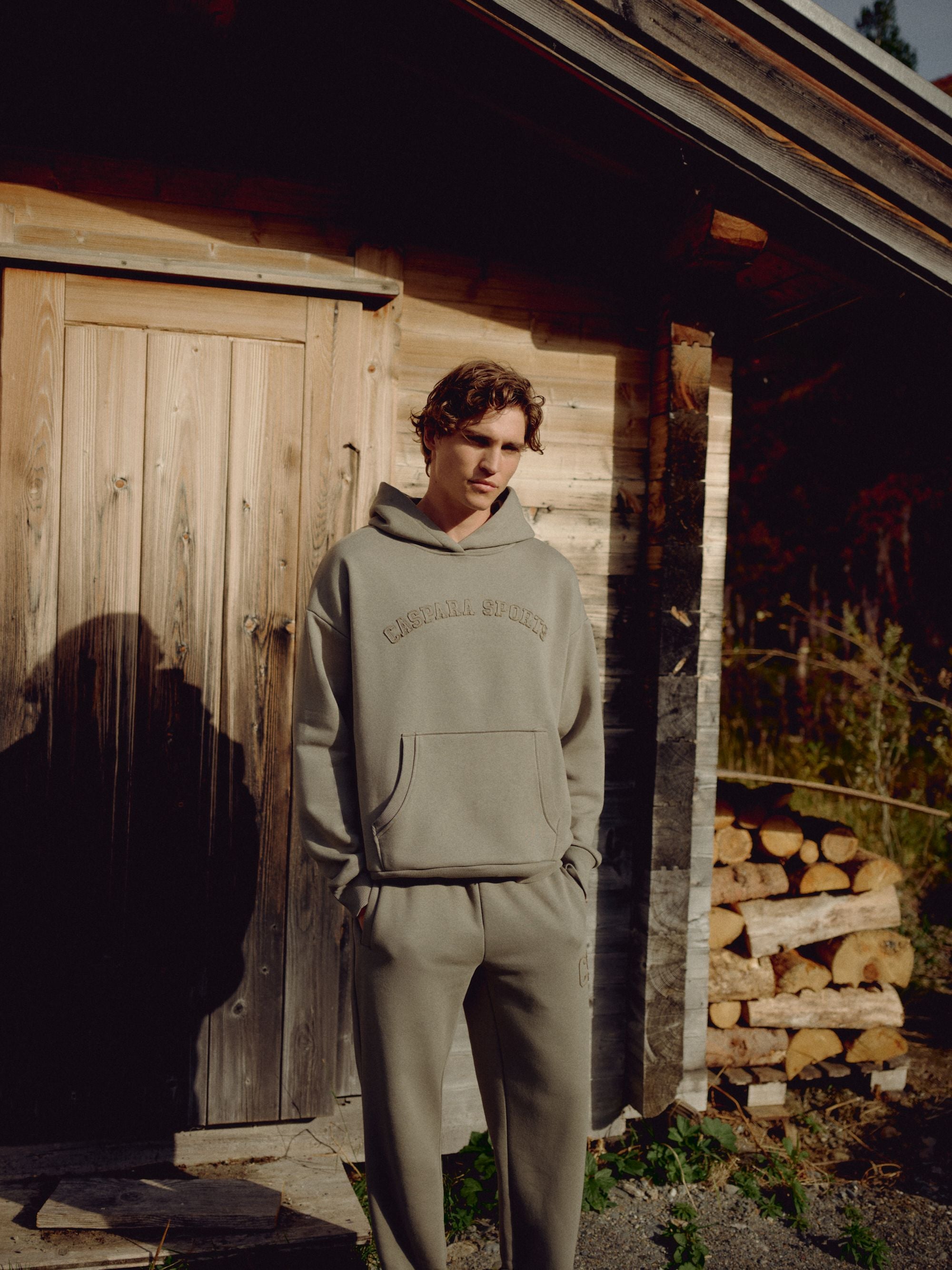 Man wearing a khaki CASPARA CS hoodie outside a rustic wooden cabin.