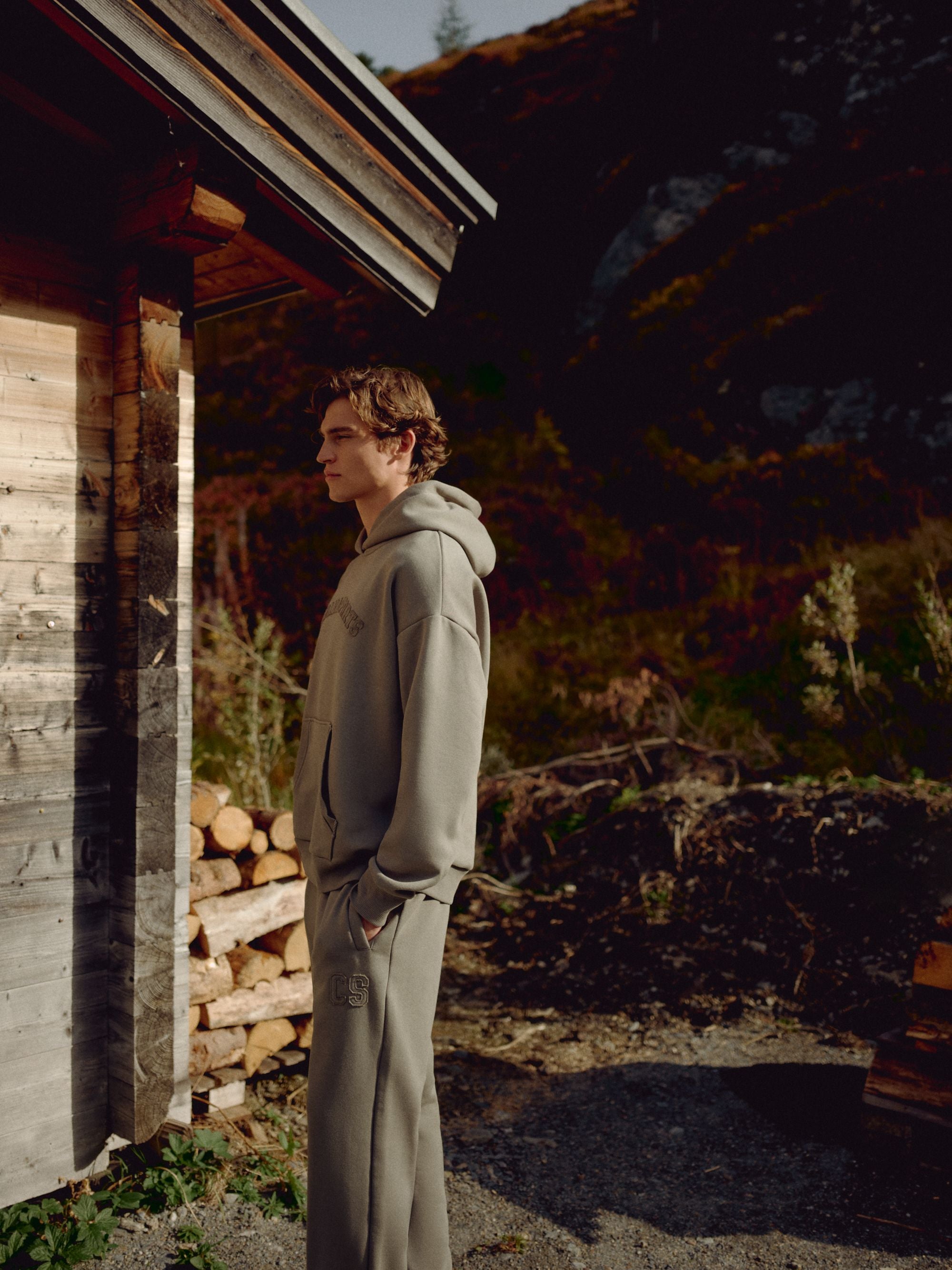 Man wearing a khaki CASPARA CS hoodie standing outdoors beside a wooden cabin.