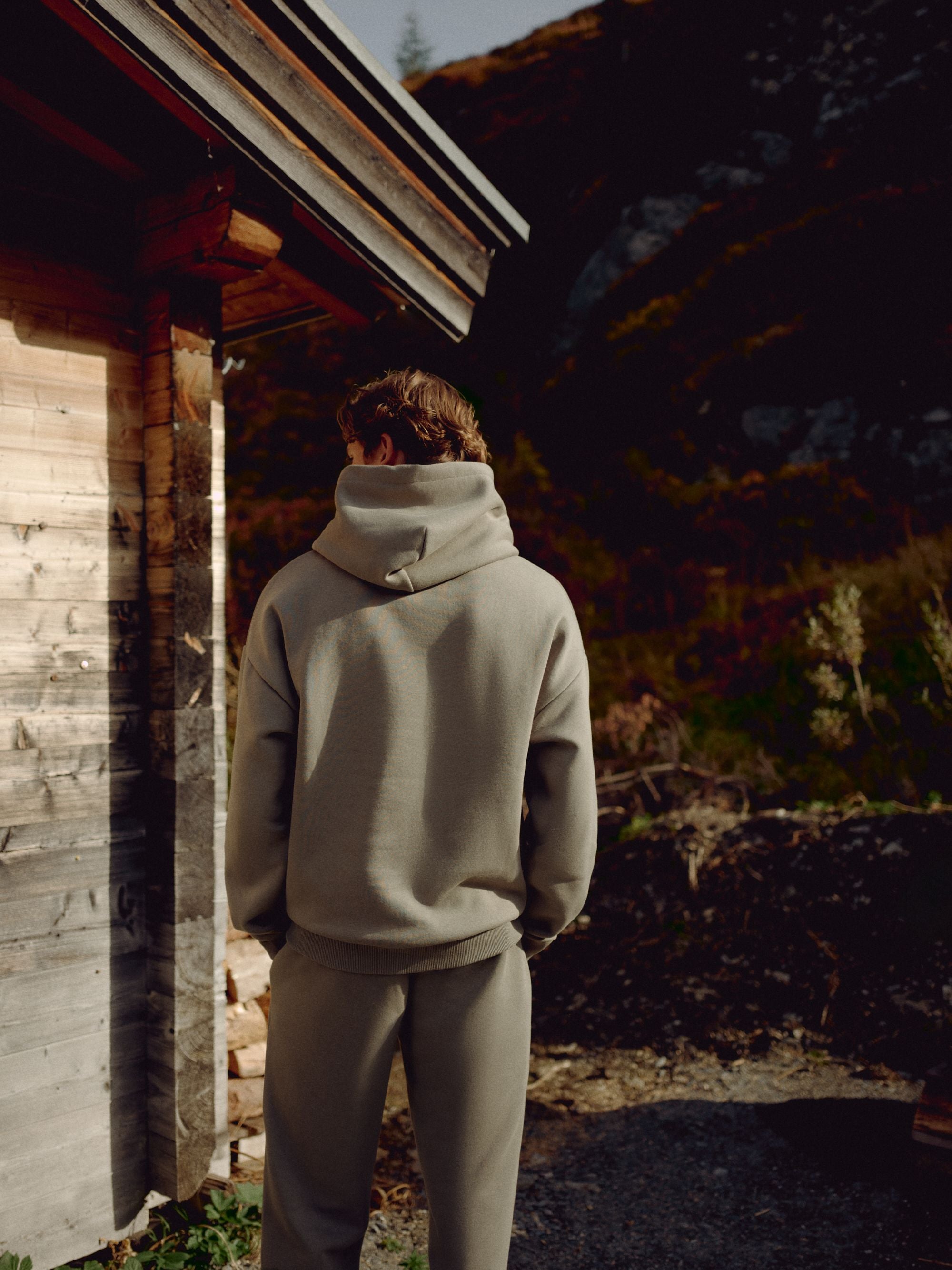 Back view of a man wearing a khaki CASPARA CS zip hoodie next to a wooden cabin.