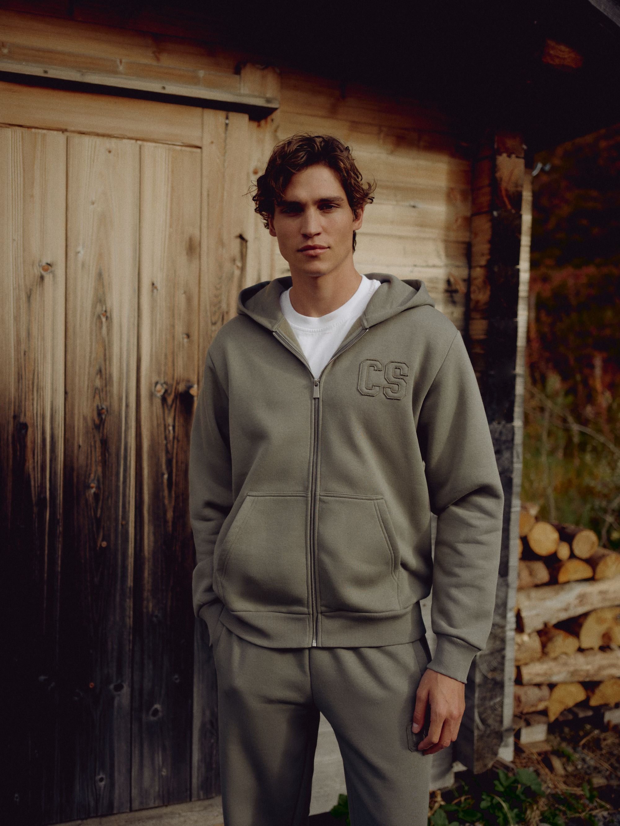 Man wearing a khaki CASPARA CS zip hoodie standing in front of a wooden cabin.