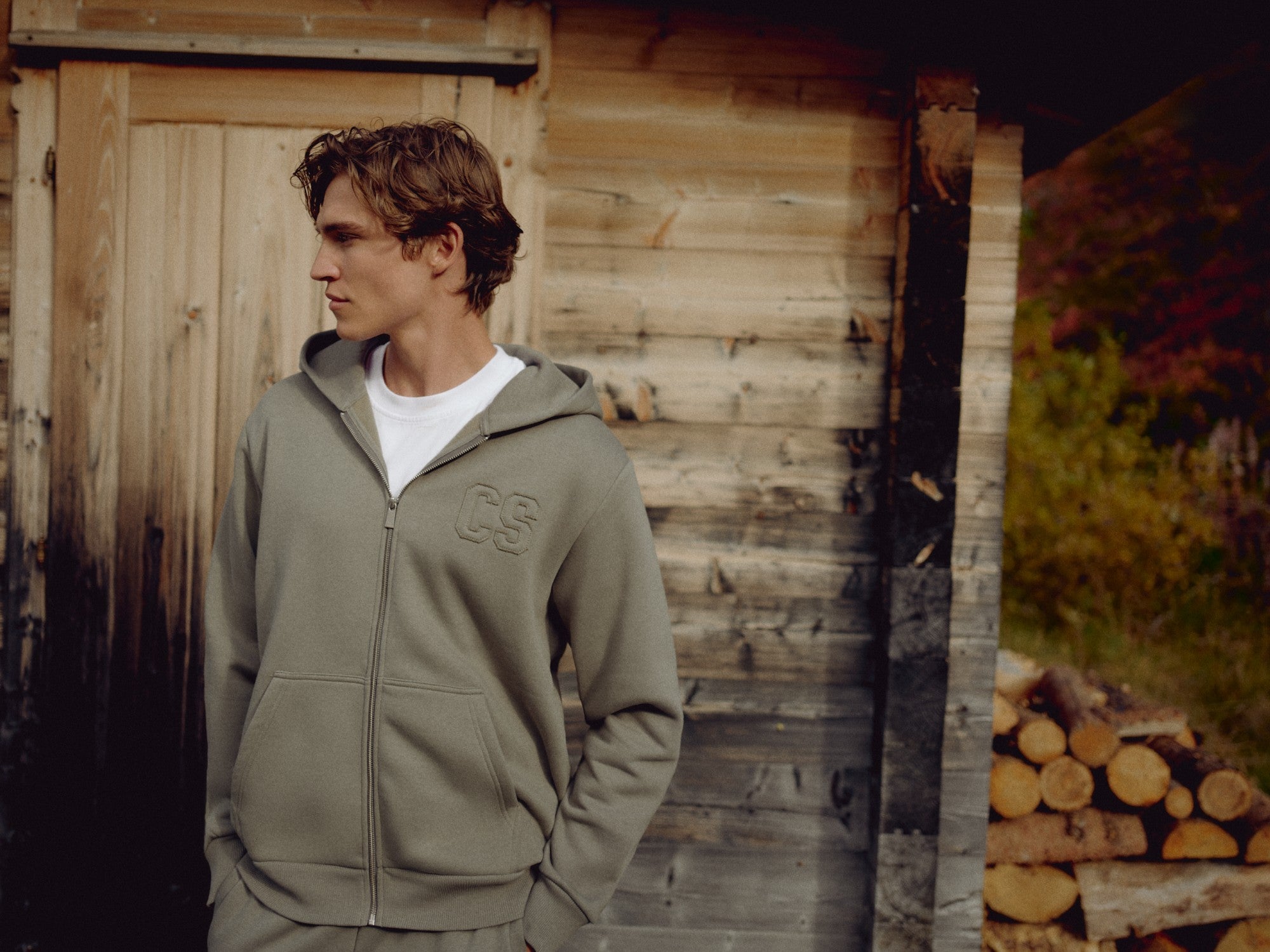 Man wearing a khaki CASPARA CS zip hoodie standing outside a rustic wooden cabin.