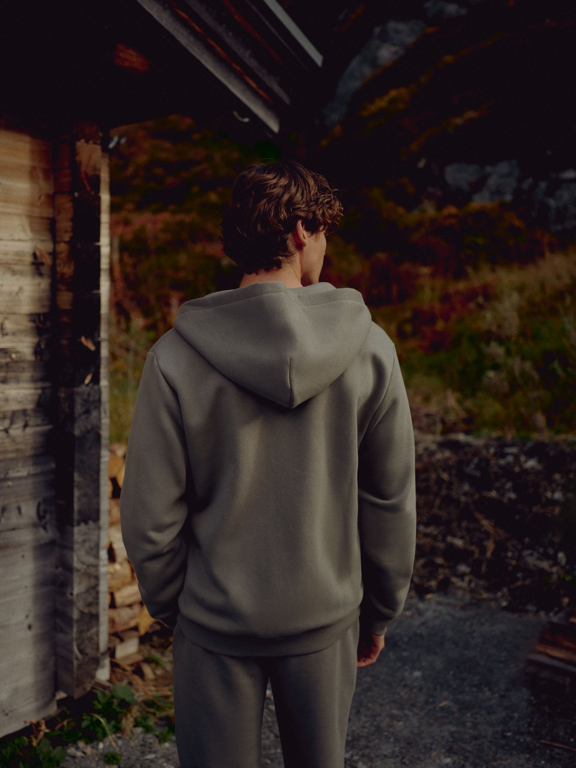 Man wearing a khaki CASPARA CS zip hoodie seen from behind outside a wooden cabin.